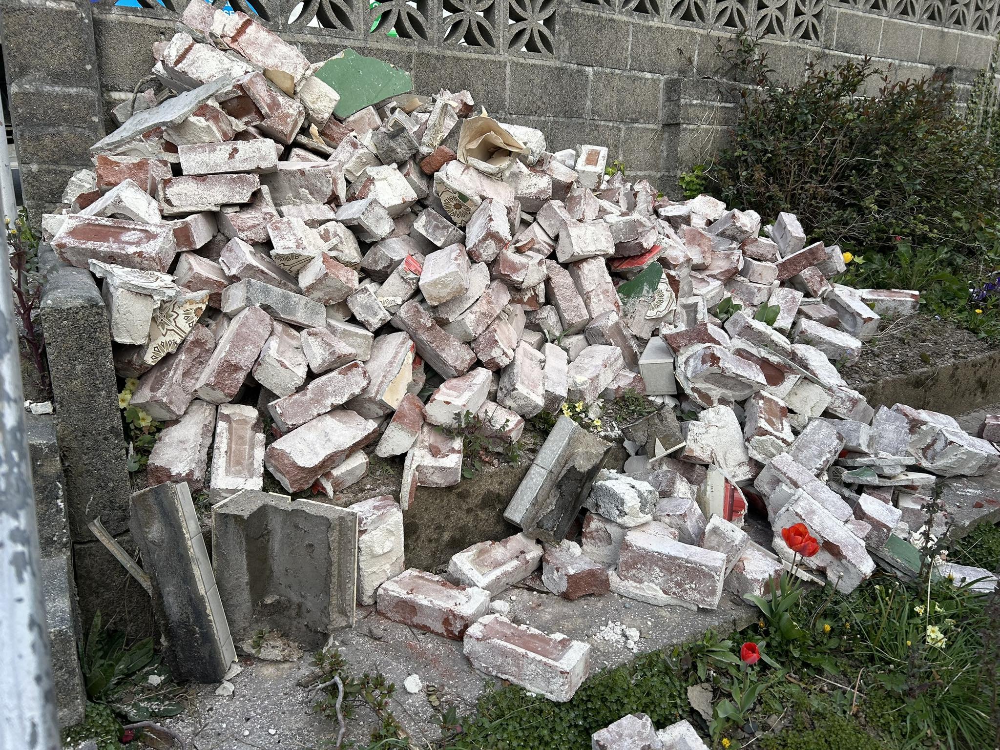 Pile of broken bricks and debris next to a concrete wall with flowers and greenery around.