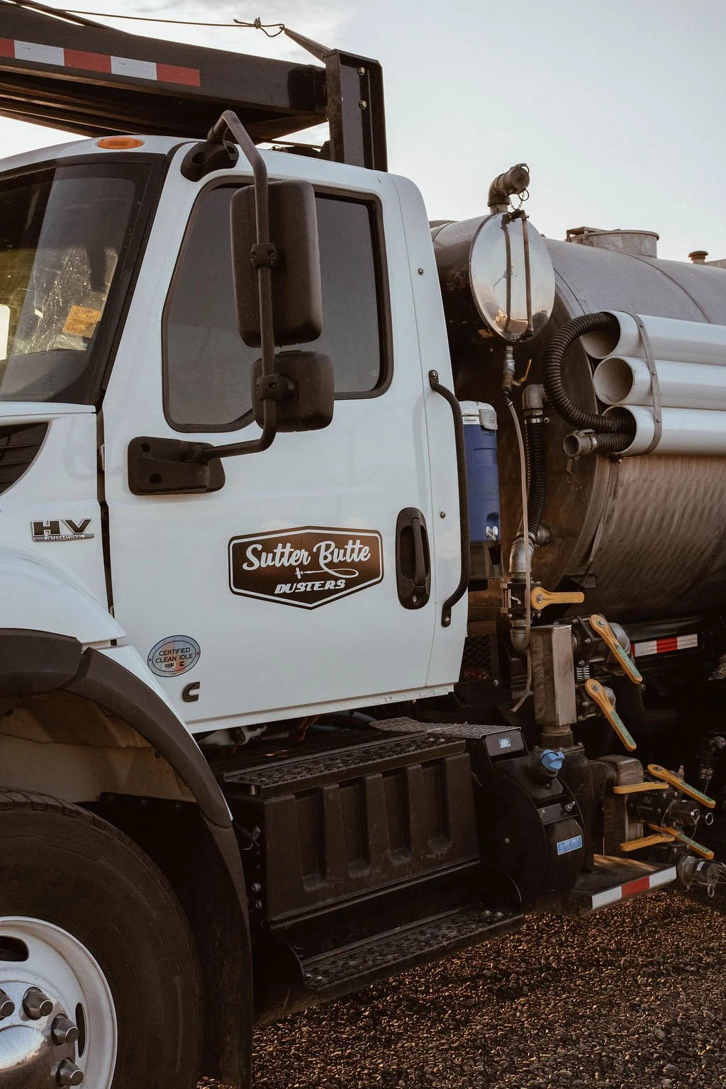 A tanker truck with the logo 'Sutter Butte Dusters' on the door, equipped with hoses and gauges, parked on gravel at sunset.