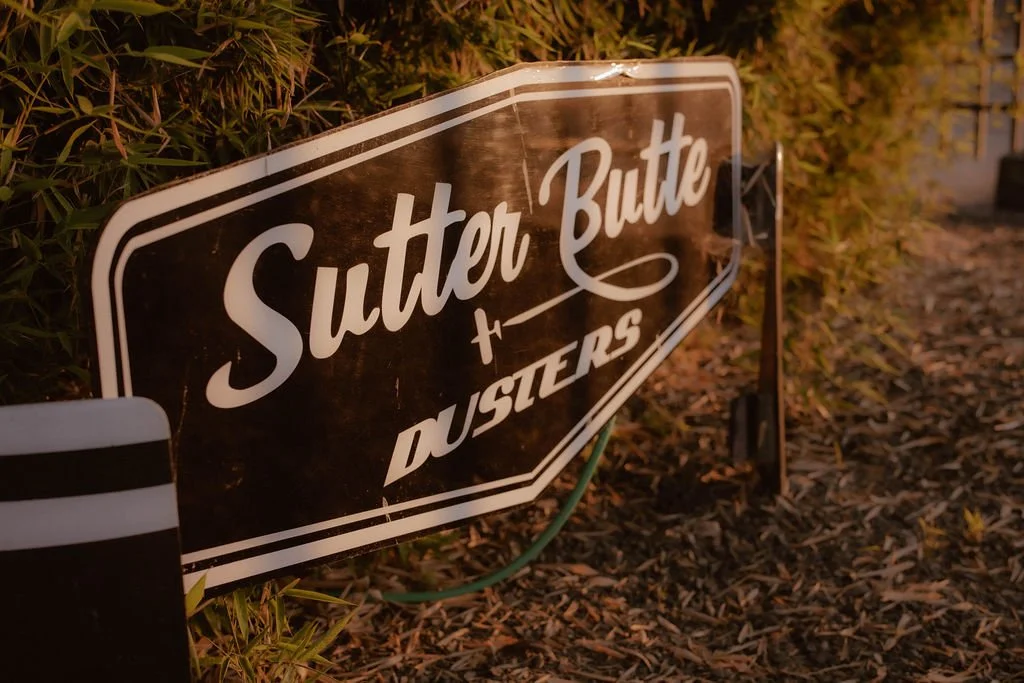 A sign for the Sutter Butte Dusters, set on a landscaped area with mulch and bushes.