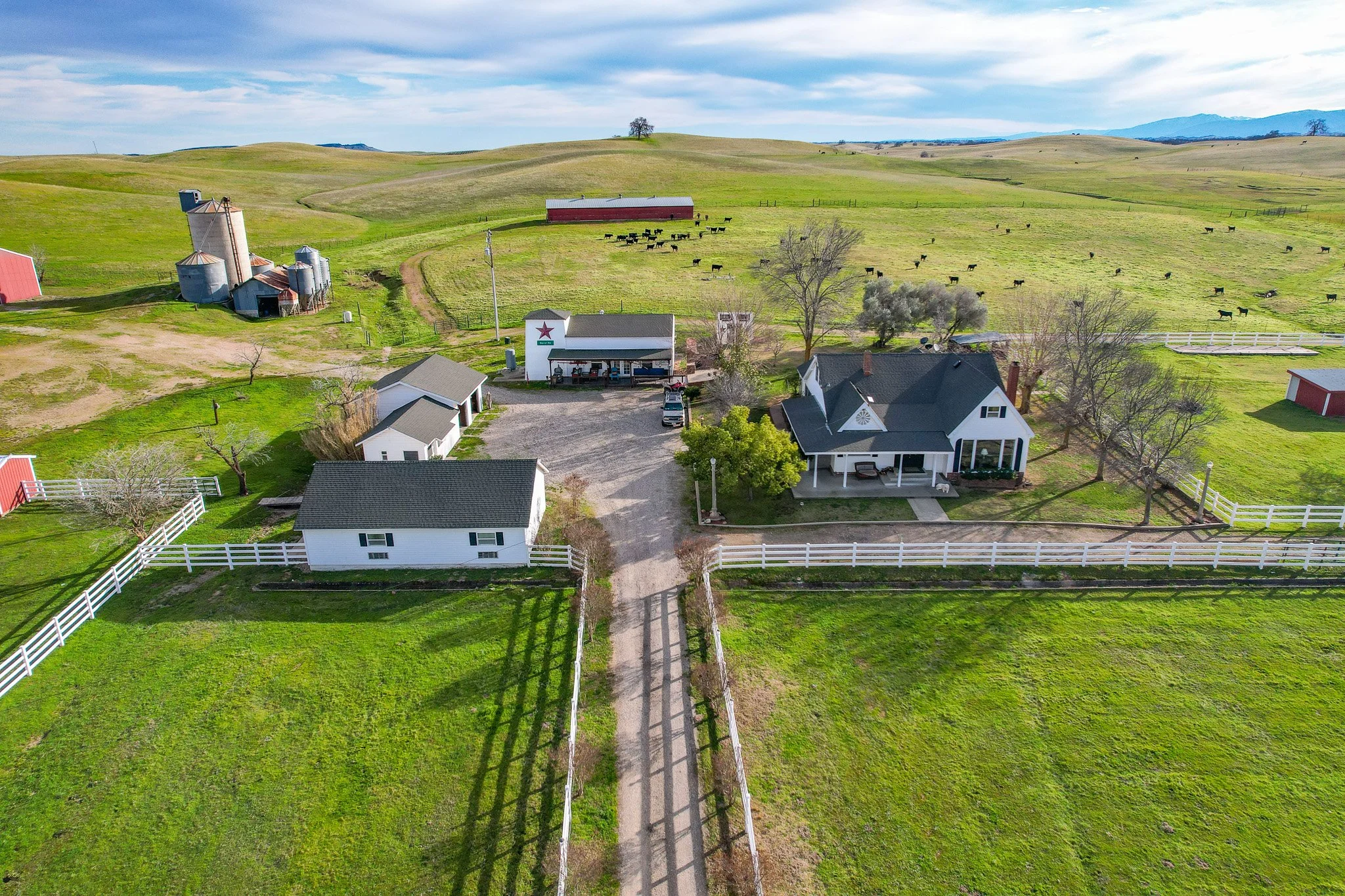 Aerial view of a rural farm with a main house, several smaller buildings, a gravel driveway, fenced green pastures, grazing cows, and rolling hills in the background.