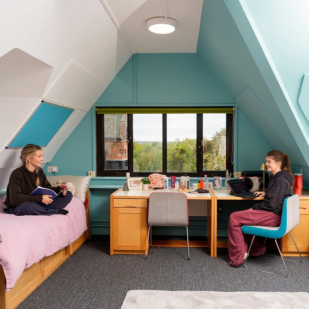 A cozy attic room with two young women smiling and engaging in conversation. One woman is sitting on a bed with a pink blanket holding a book, and the other is seated at a desk with a laptop. The room has a large window with a view of trees and houses outside, and various personal items and toiletries on the desk.