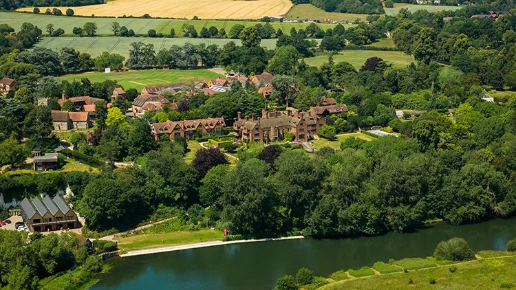 Aerial view of a river lined with trees and houses in a lush green landscape with rolling hills and farmland in the background.