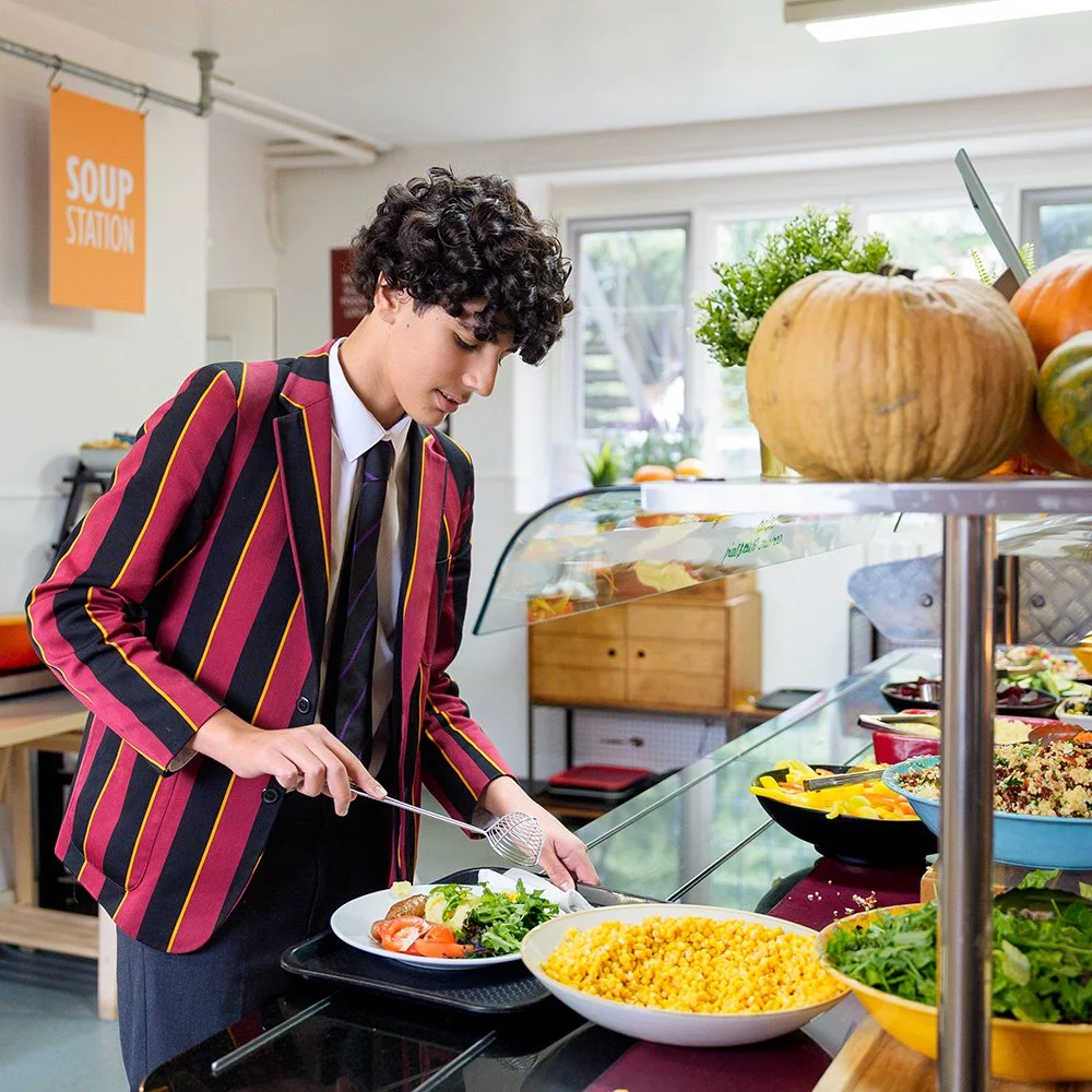 A young man in a maroon, black, and yellow striped blazer serving himself salad at a cafeteria salad bar with various bowls of food including corn and greens, with a pumpkin and other vegetables on the counter.