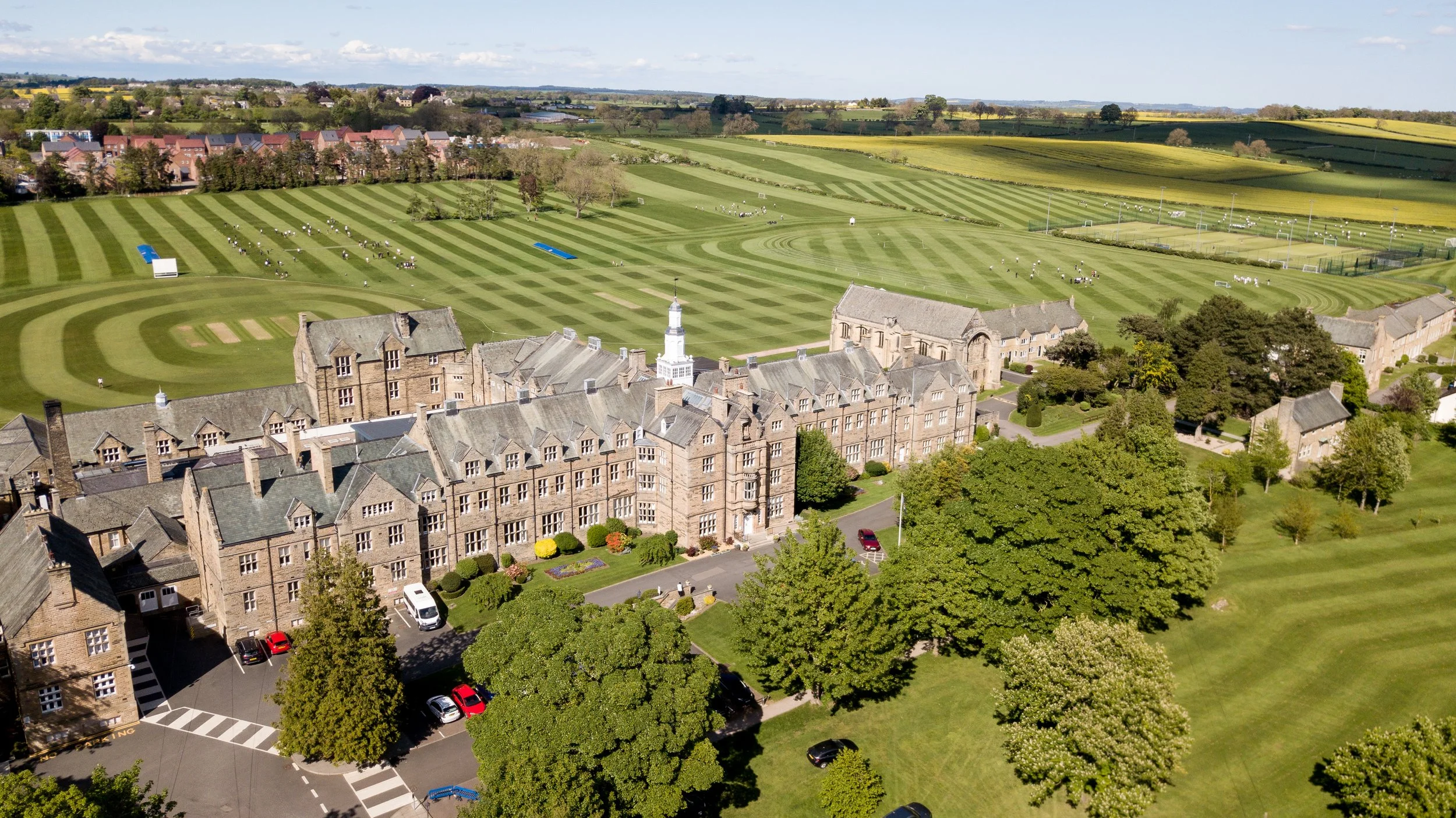 Aerial view of a large historic building with a clock tower, surrounded by a beautifully maintained lawn, trees, and sports fields in a rural area.