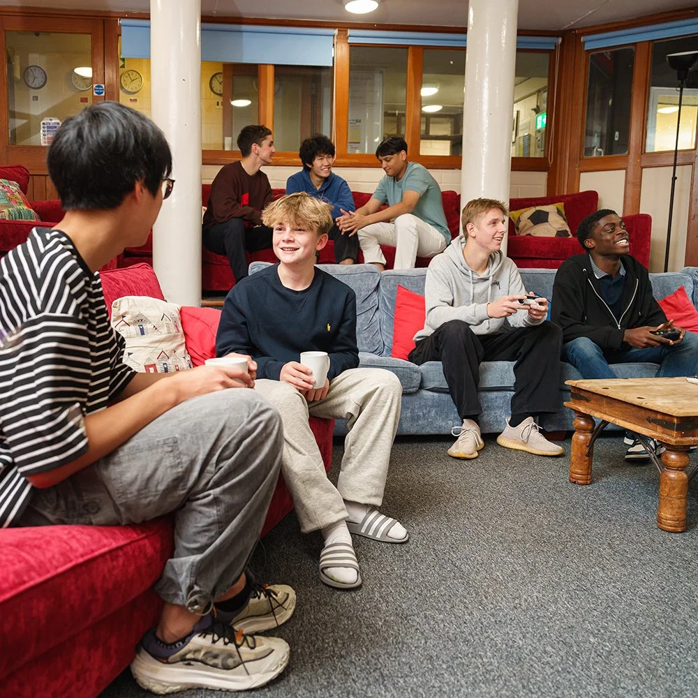 Group of young men sitting on couches in a lounge, some holding cups and gaming controllers, engaging in conversation and enjoying each other's company.