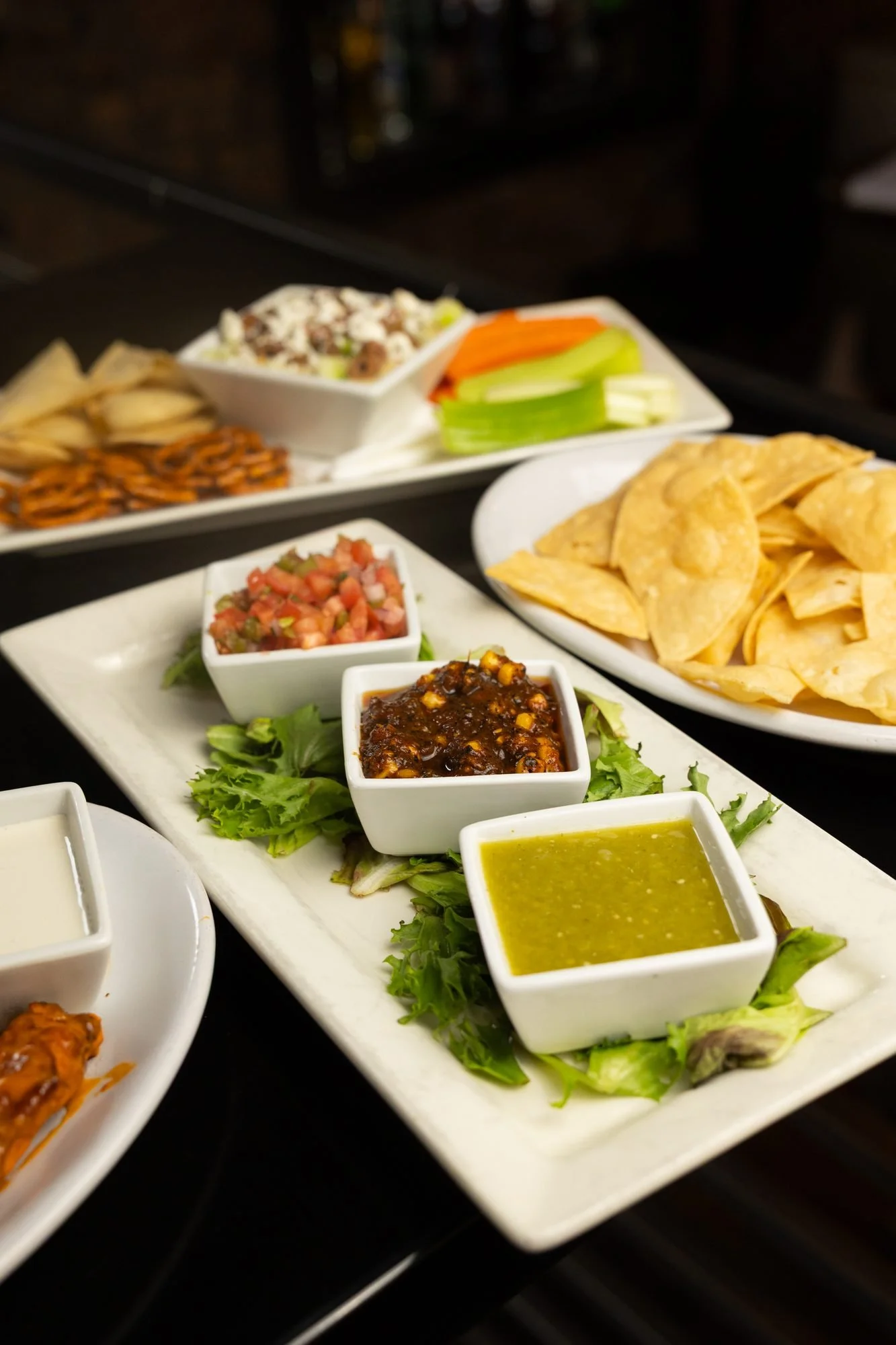 Several appetizer plates spread across a dark table with chips, salsa, dips, wings, and greens