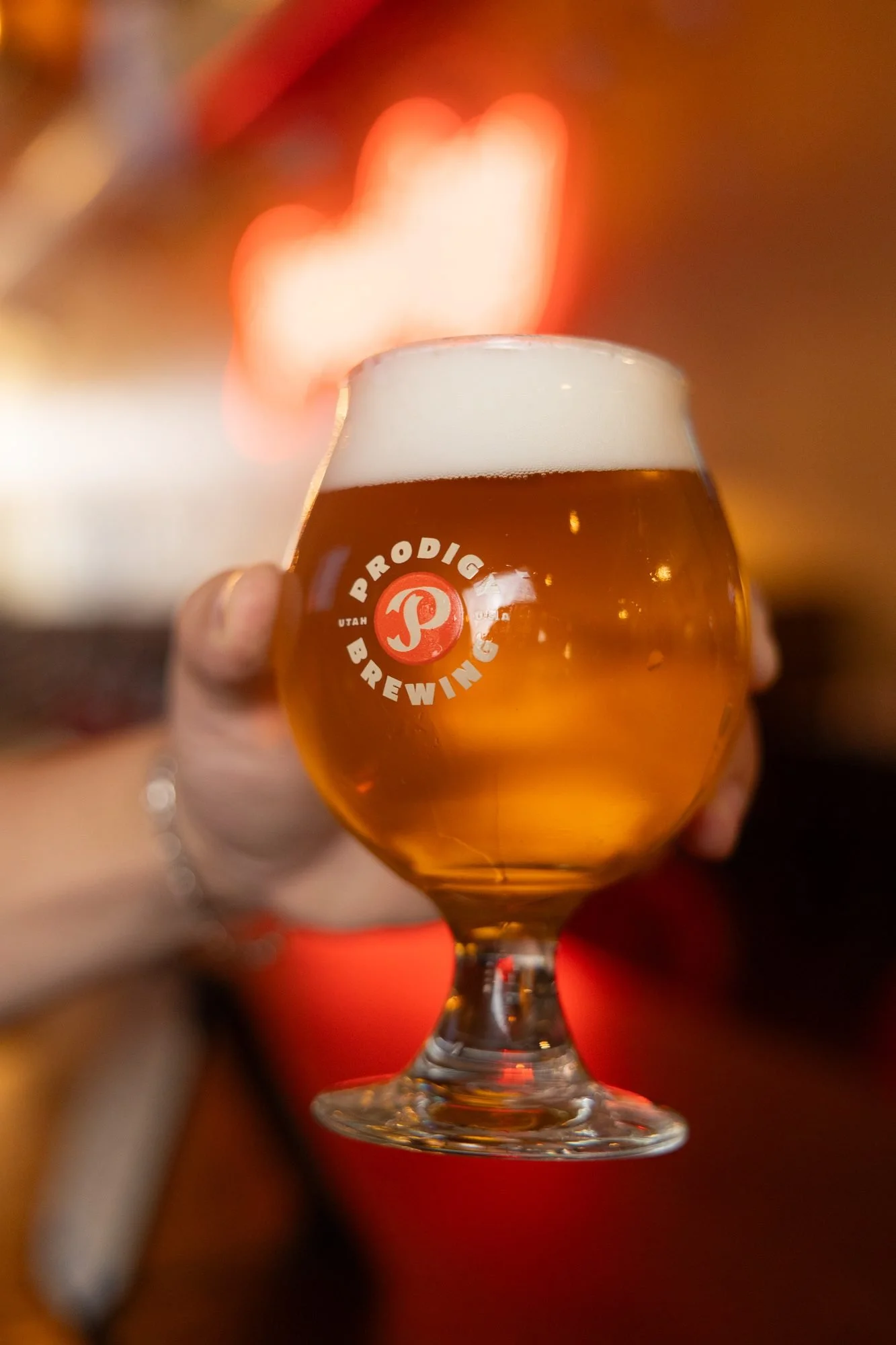 Glass of amber beer raised in front of the red neon sign at Prodigy Brewing in Logan