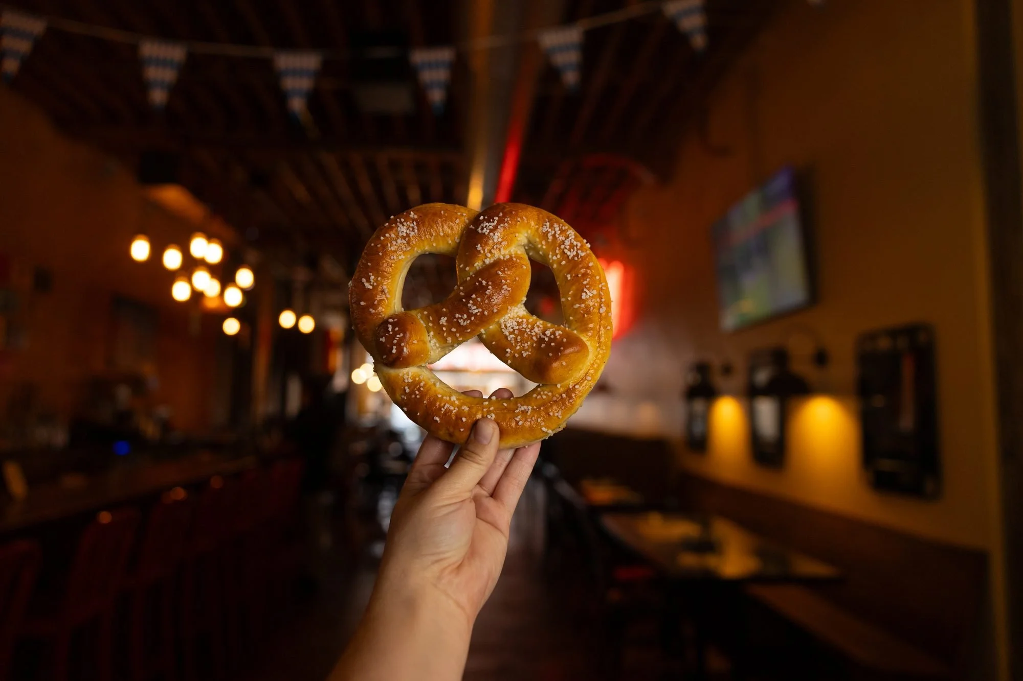 A hand holds a salted soft pretzel up in a dark dining room strung with small lights