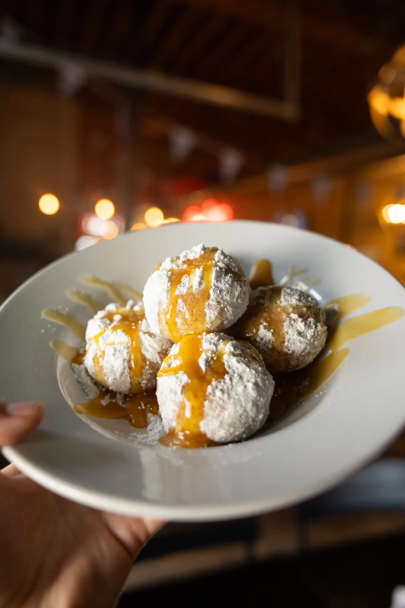A hand holds a white plate of powdered sugar dough balls drizzled with caramel in a dim dining room
