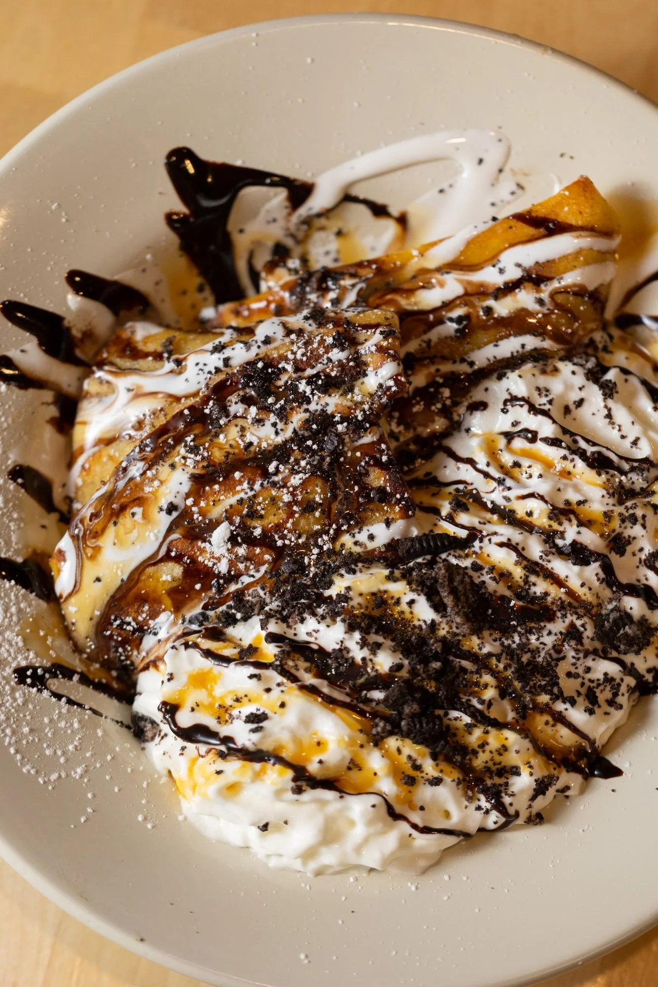 A folded crepe on a white plate covered in whipped cream, chocolate drizzle and crushed cookie pieces