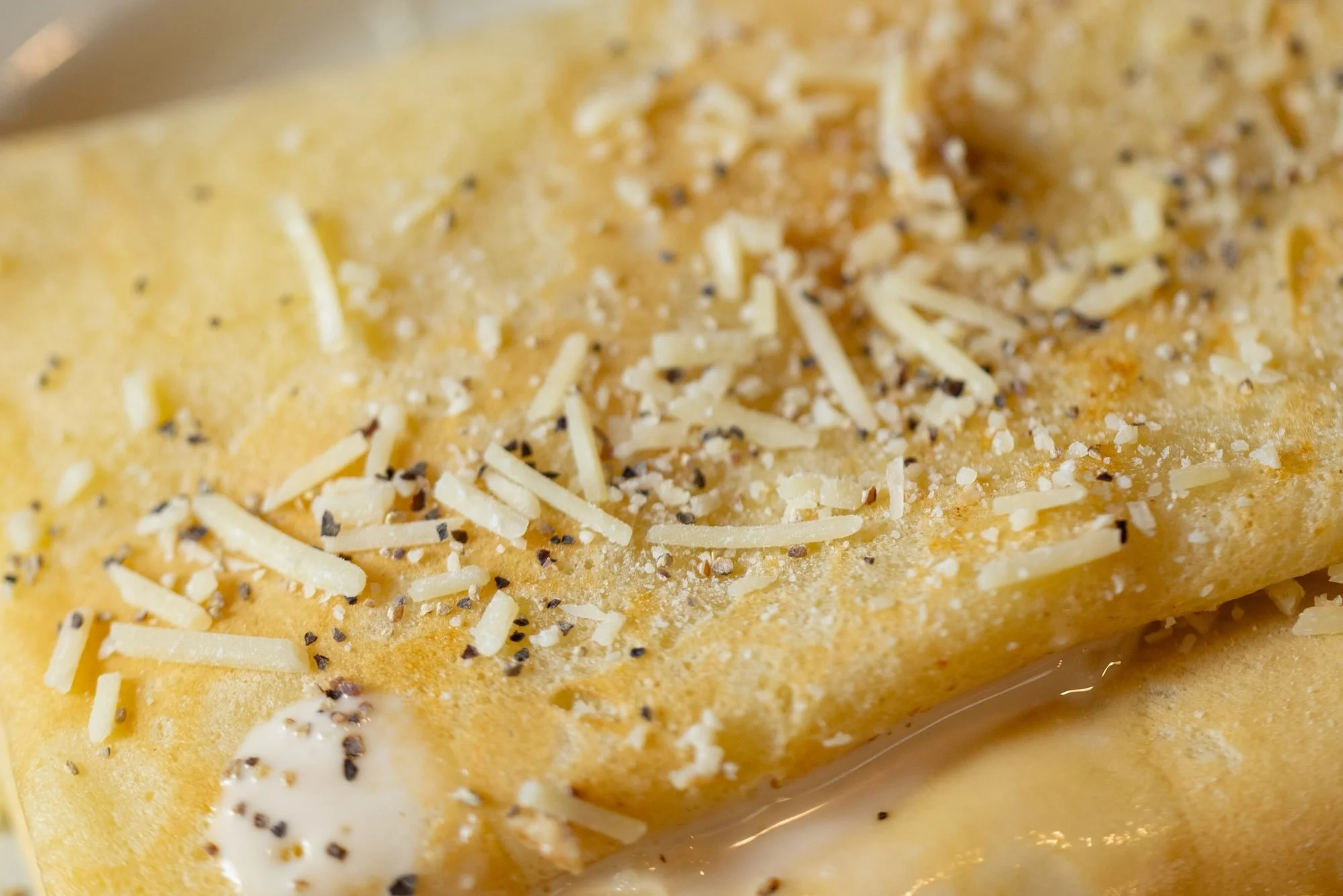 Close view of a folded golden crepe topped with shredded parmesan cheese and black pepper on a white plate