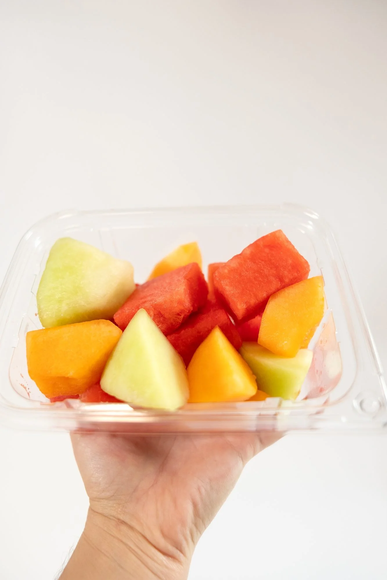 A hand holds a clear container of cut watermelon, cantaloupe, and honeydew up against a white background
