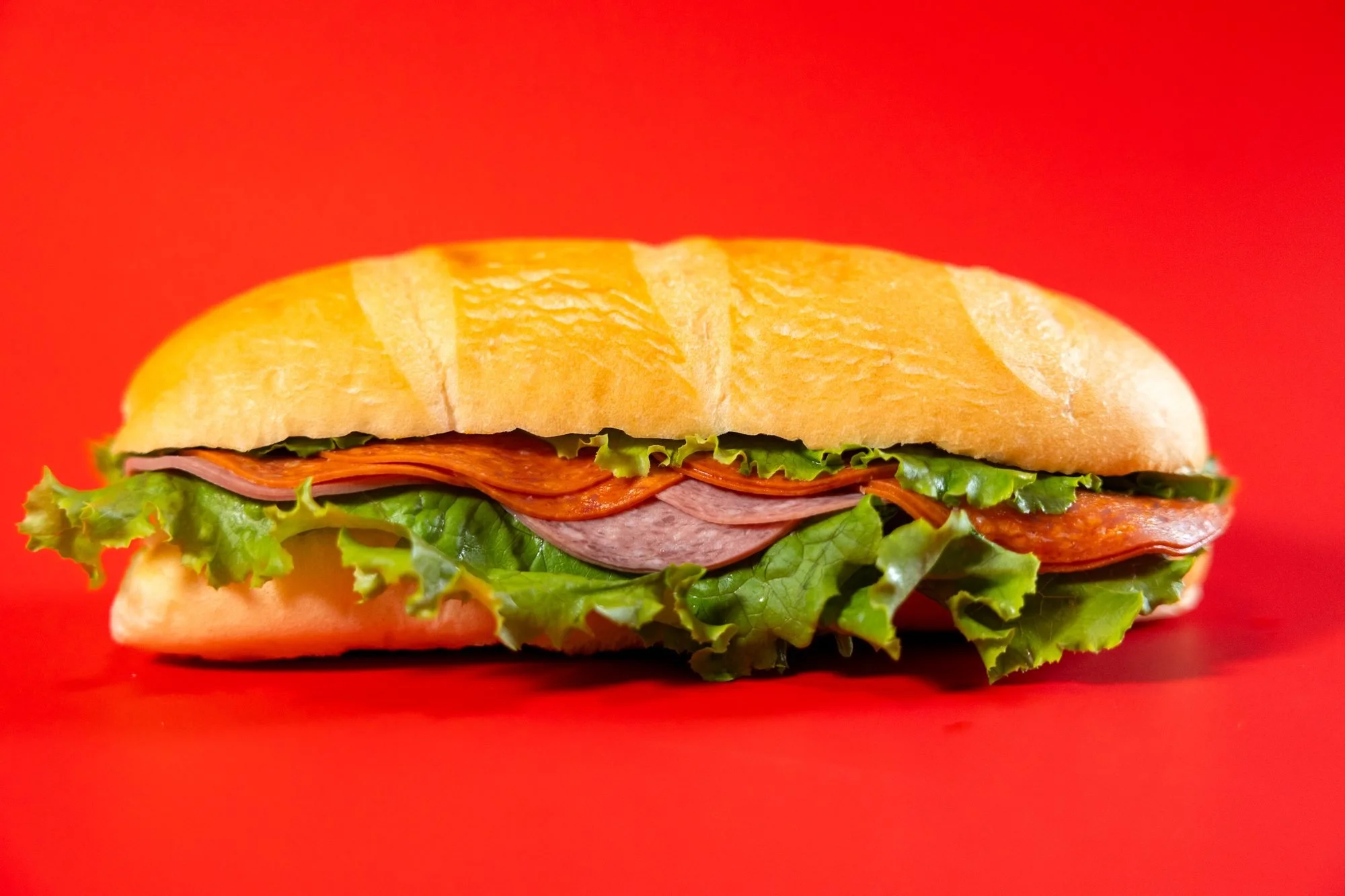 A sub sandwich with deli meat, lettuce, and tomato sits on a solid red background