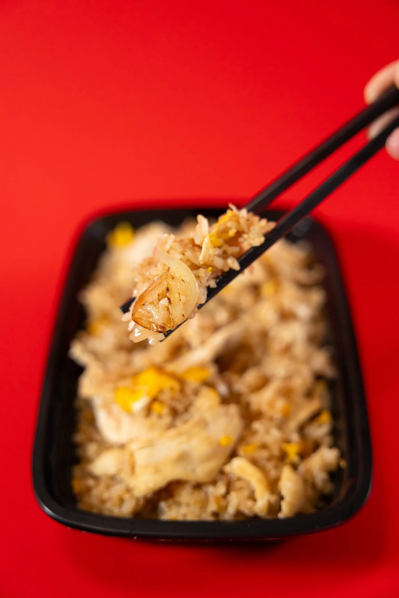Chopsticks lift a piece of chicken from a black container of fried rice on a red background