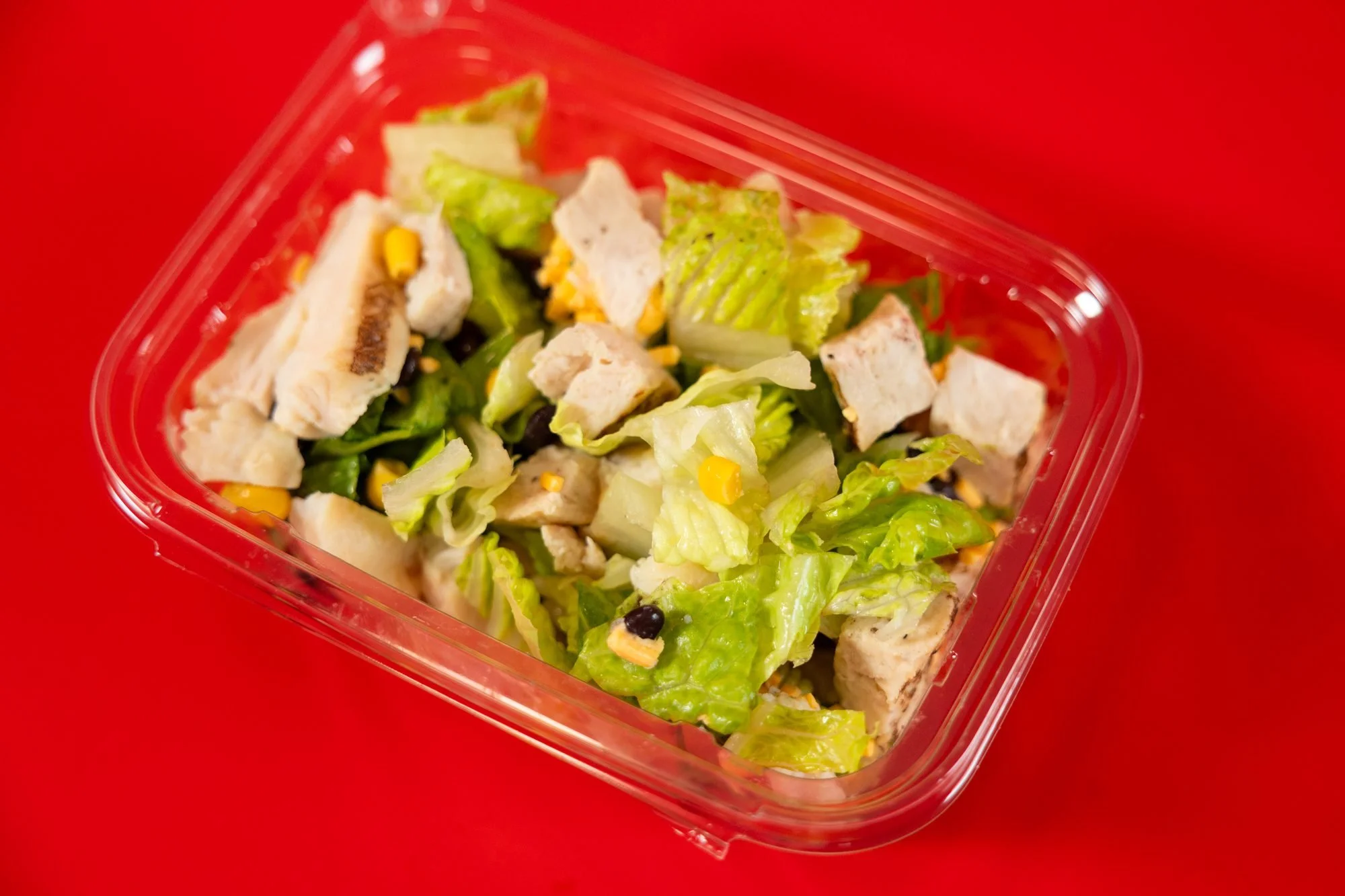 A clear container of chopped chicken over romaine lettuce with corn sits on a red background