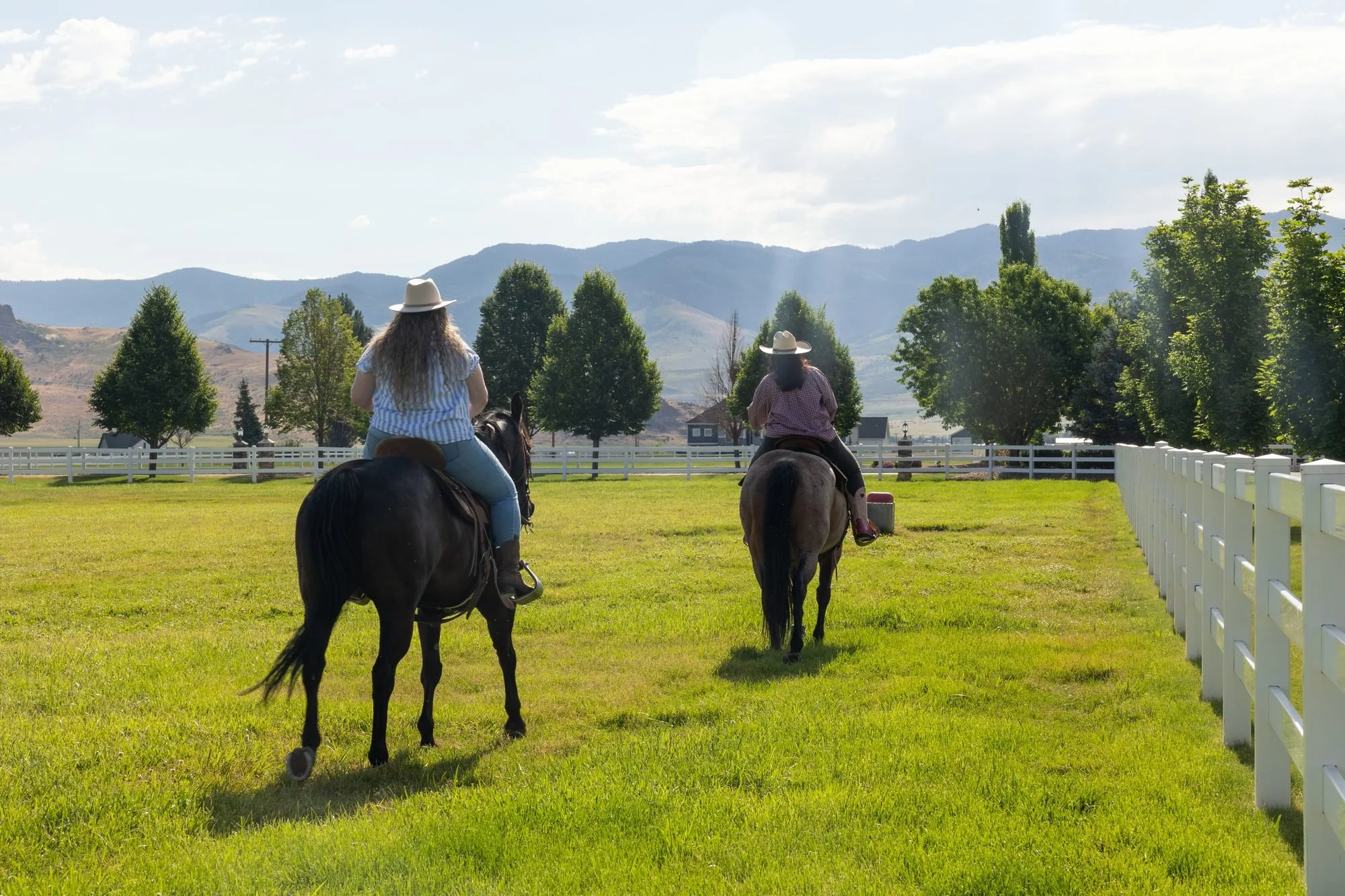 Two riders on horseback cross a green pasture beside a white fence with mountains and clouds behind