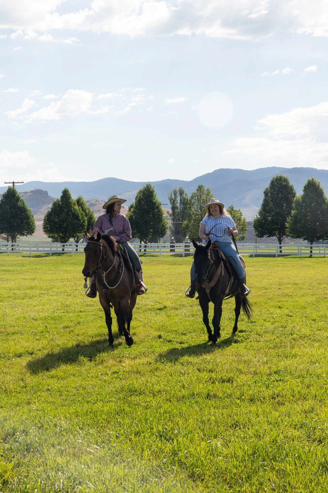 Two people on horseback ride away across a green field toward trees and mountains under a partly cloudy sky