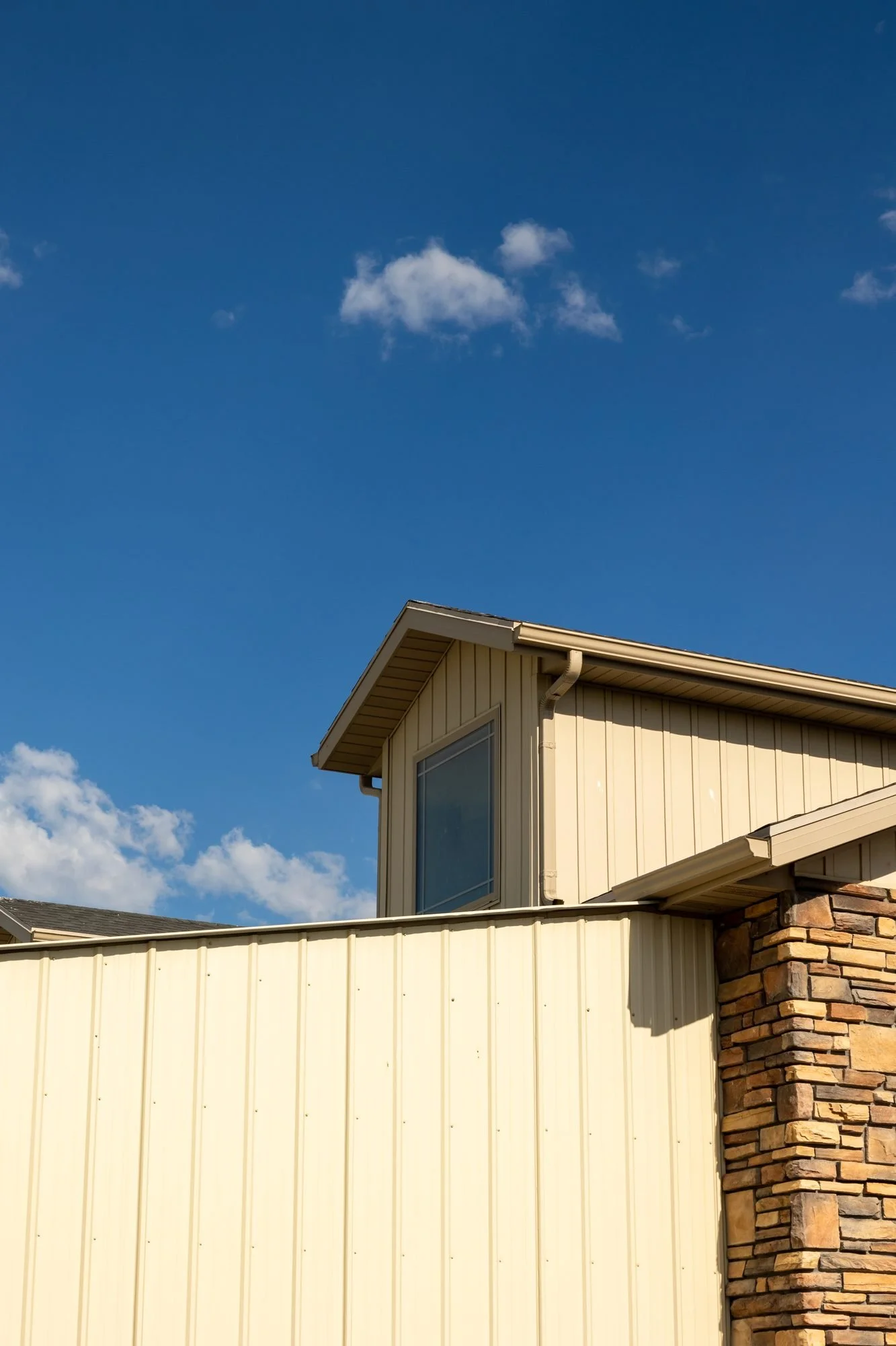 The eave of a tan barn roof and a stone-clad wall against a deep blue sky with a small cloud