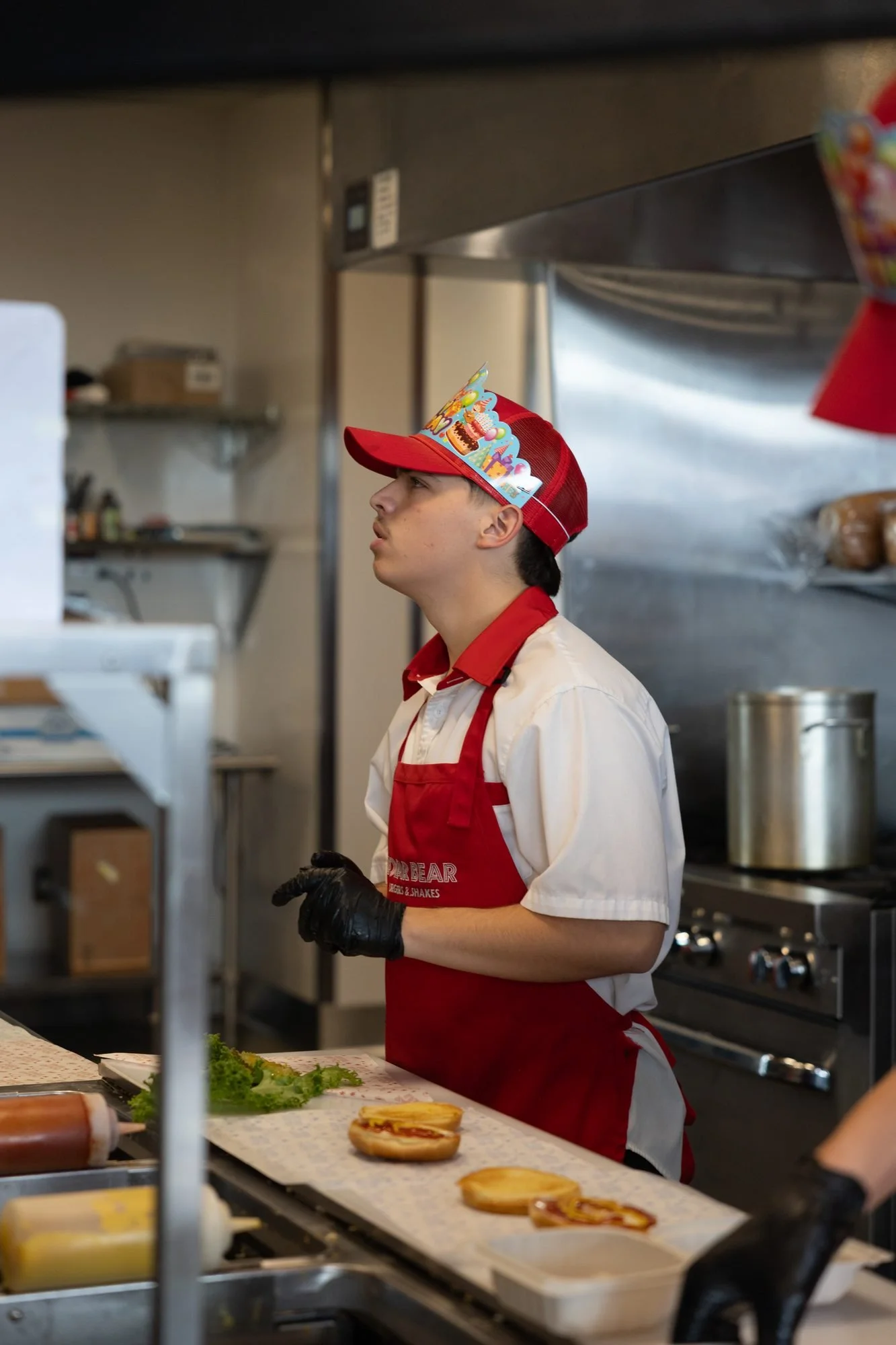Employee in a white shirt, red apron and birthday hat working the grill line inside the kitchen