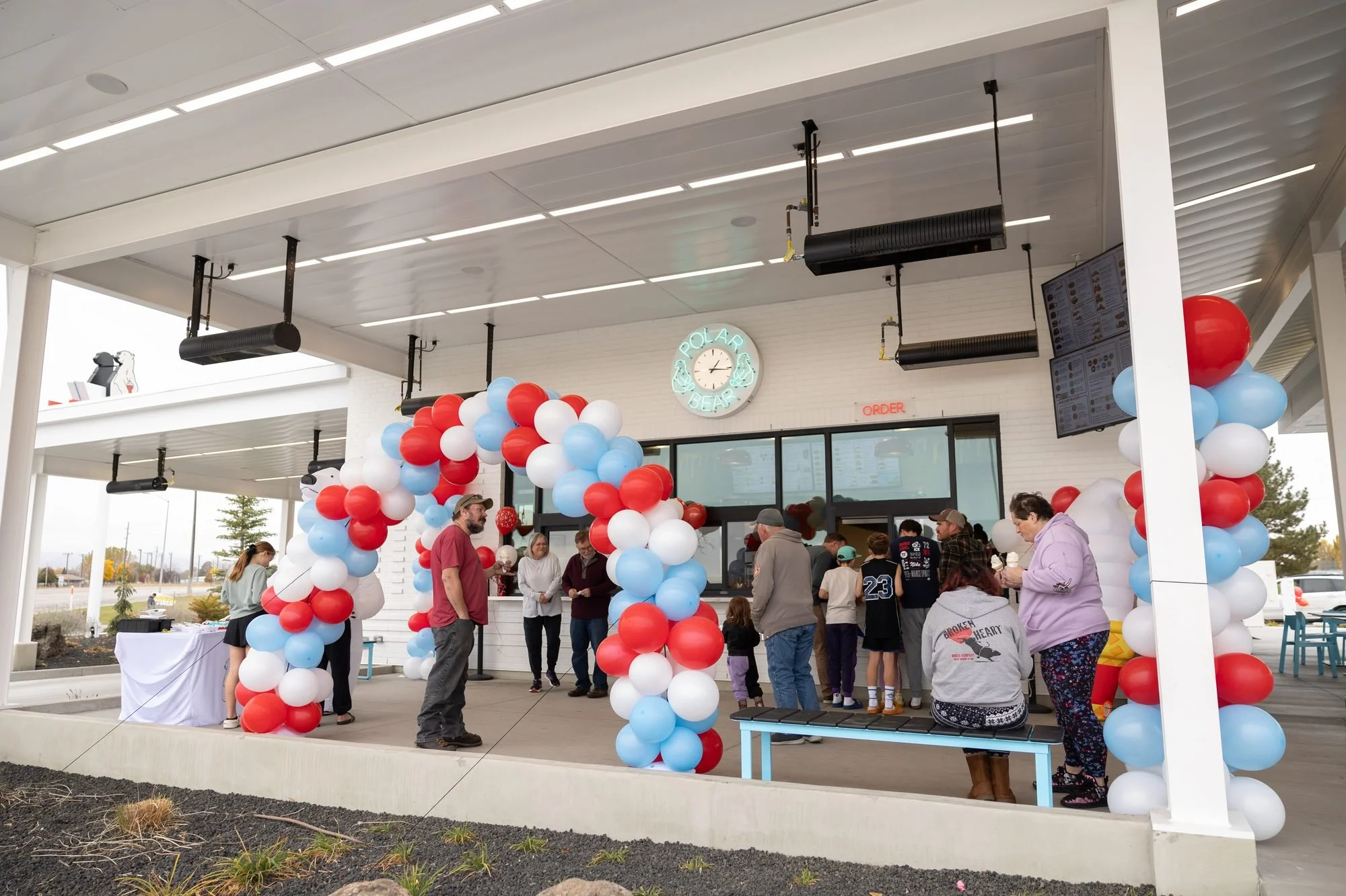 Crowd of families gathered under the covered patio beside a red, white and blue balloon arch