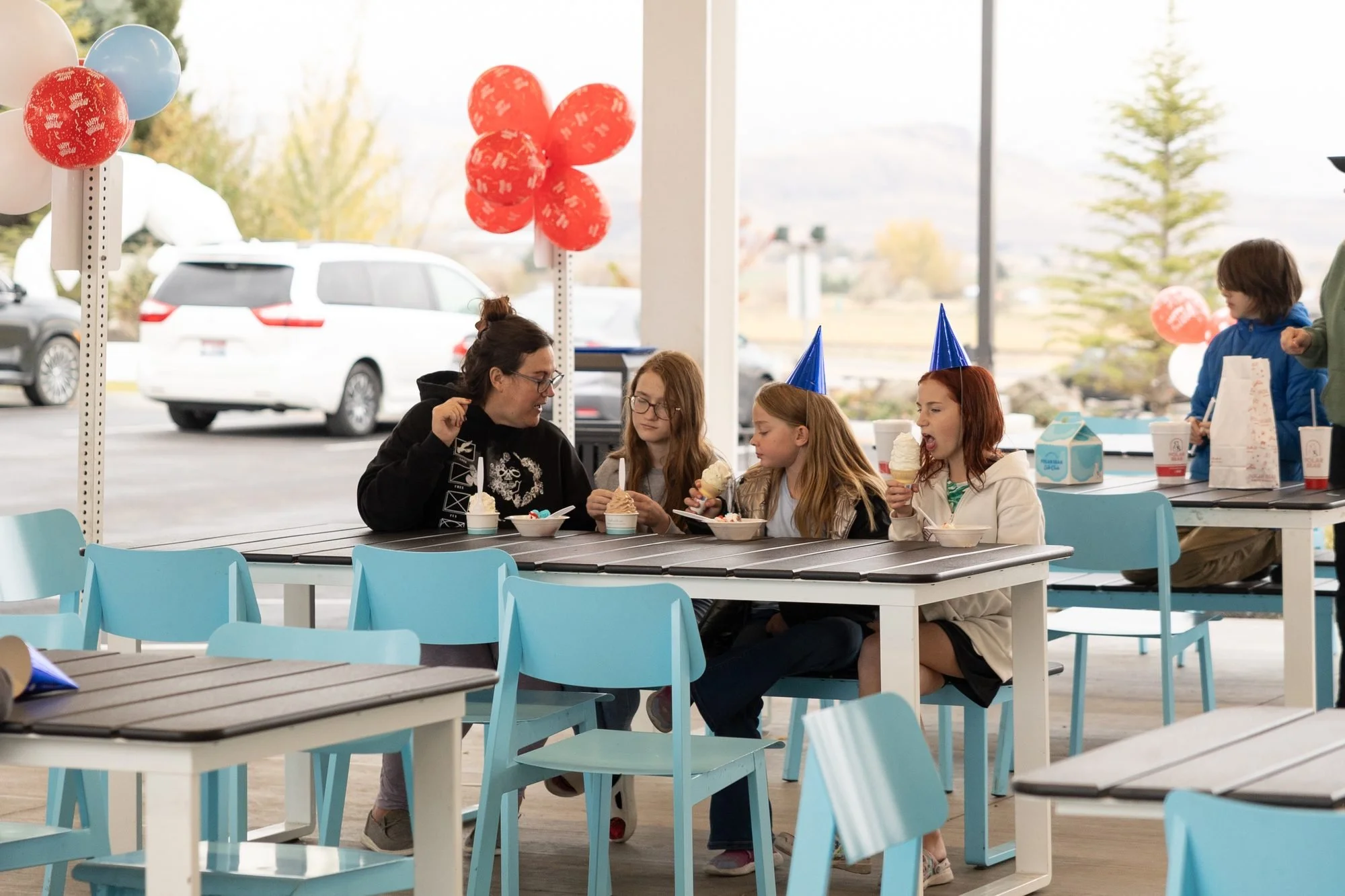 Group of girls in paper party hats sitting at light blue picnic tables with balloons and cars behind them