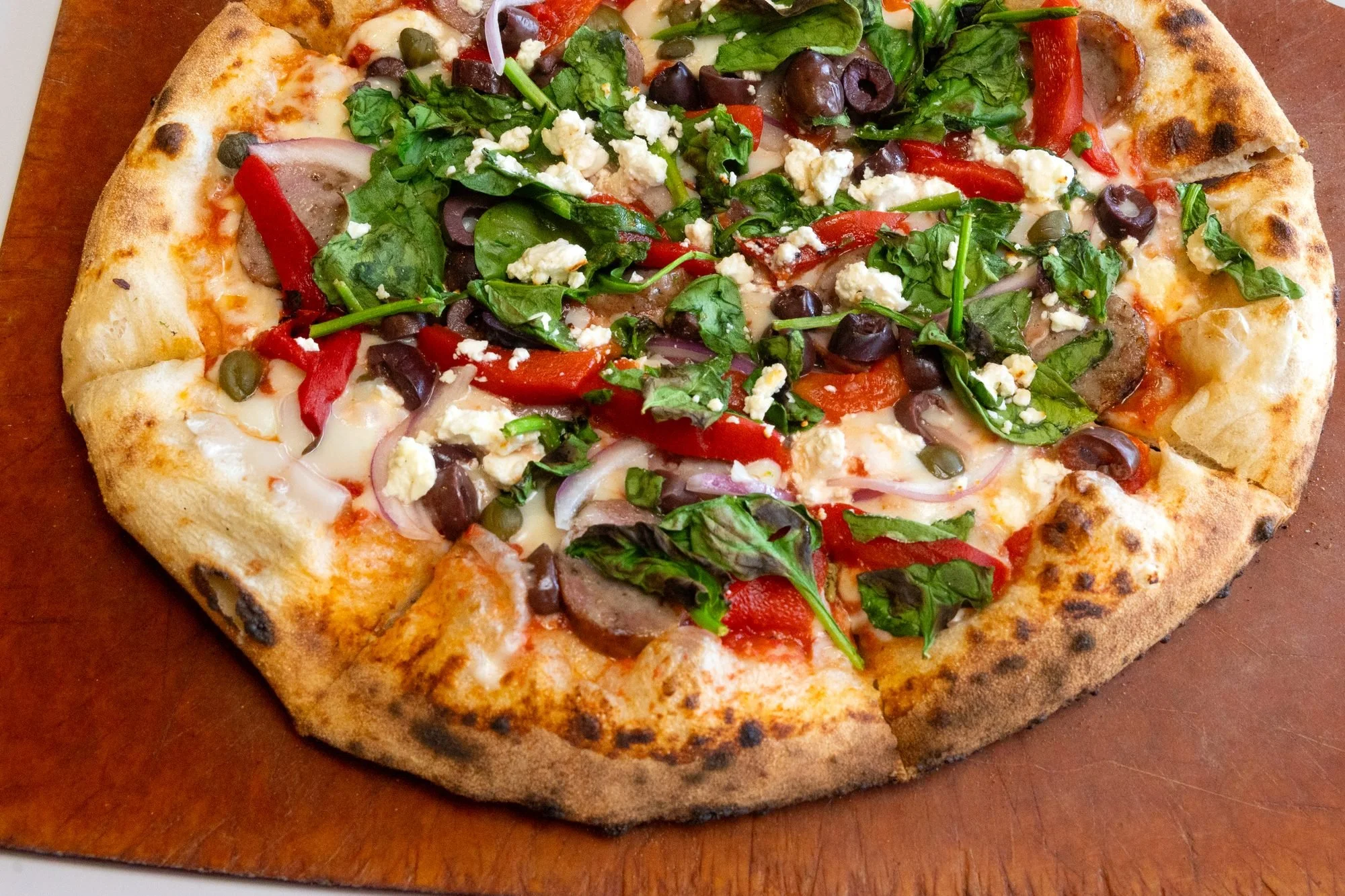 A wood-fired pizza topped with spinach, roasted red peppers, olives and feta on a wooden board