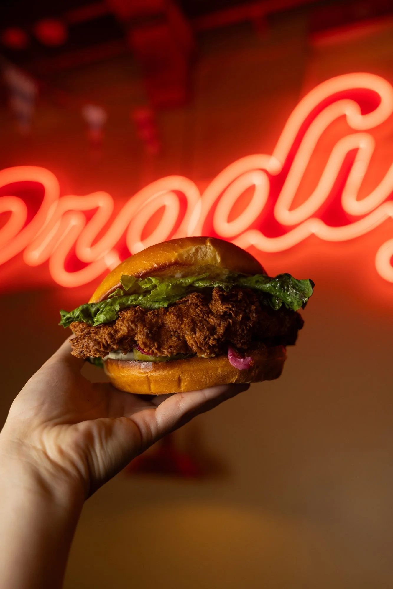 A hand holds up a fried chicken sandwich in front of a glowing red neon Prodigy sign