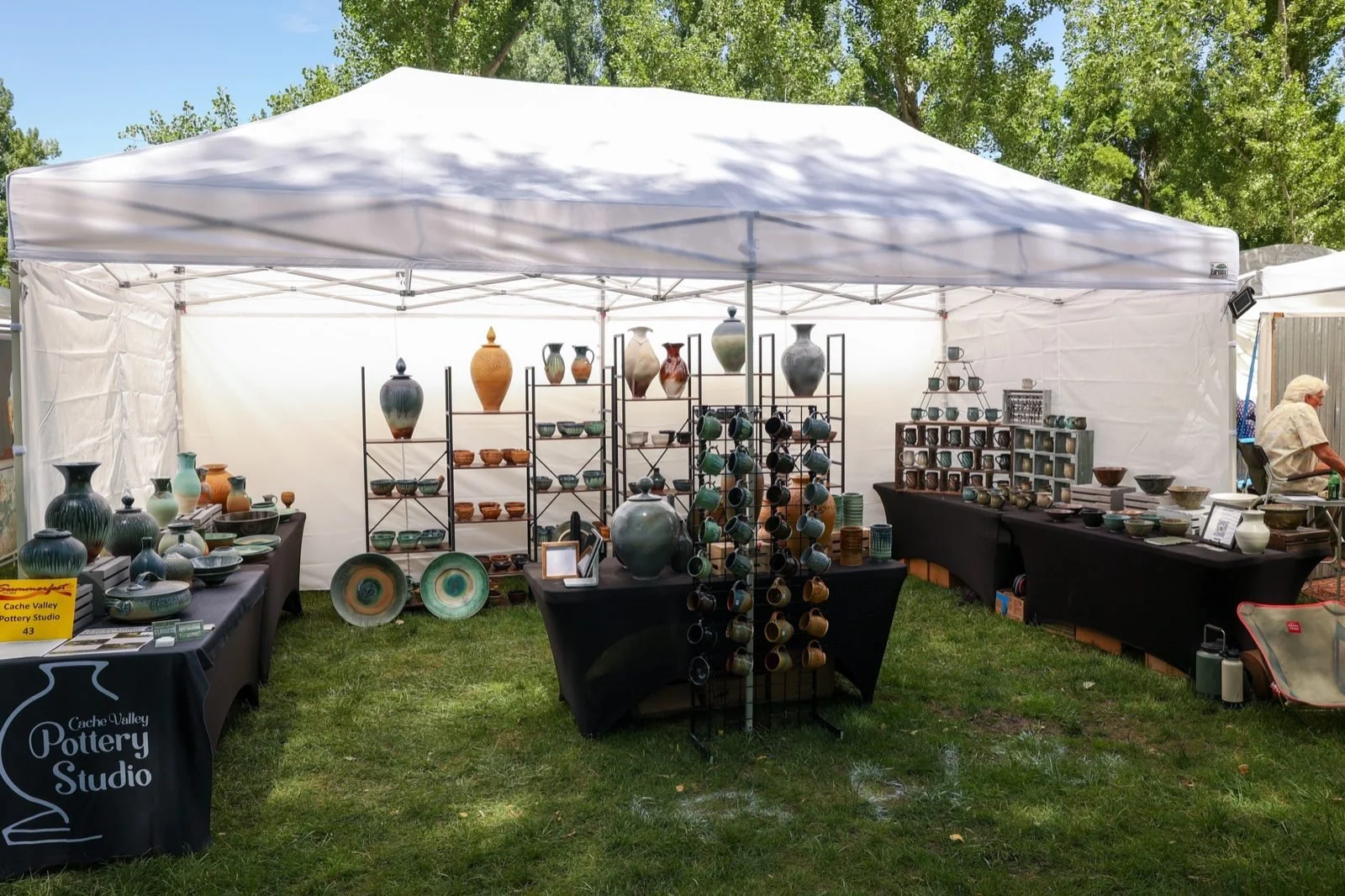 A white canopy booth of pottery set up on a shaded lawn under tall trees