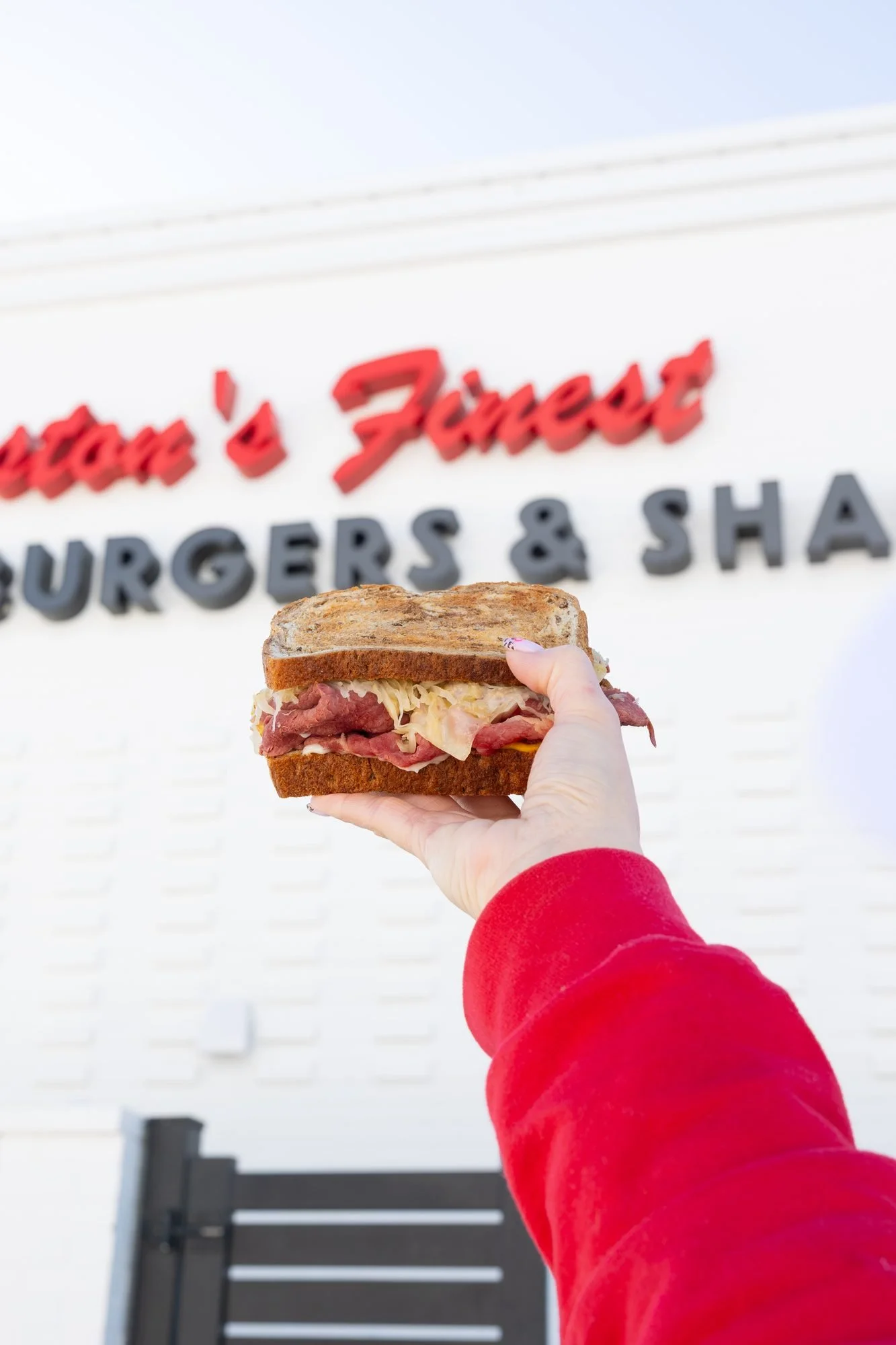 A hand holds a sandwich up in front of a white wall lettered Preston's Finest Burgers and Shakes