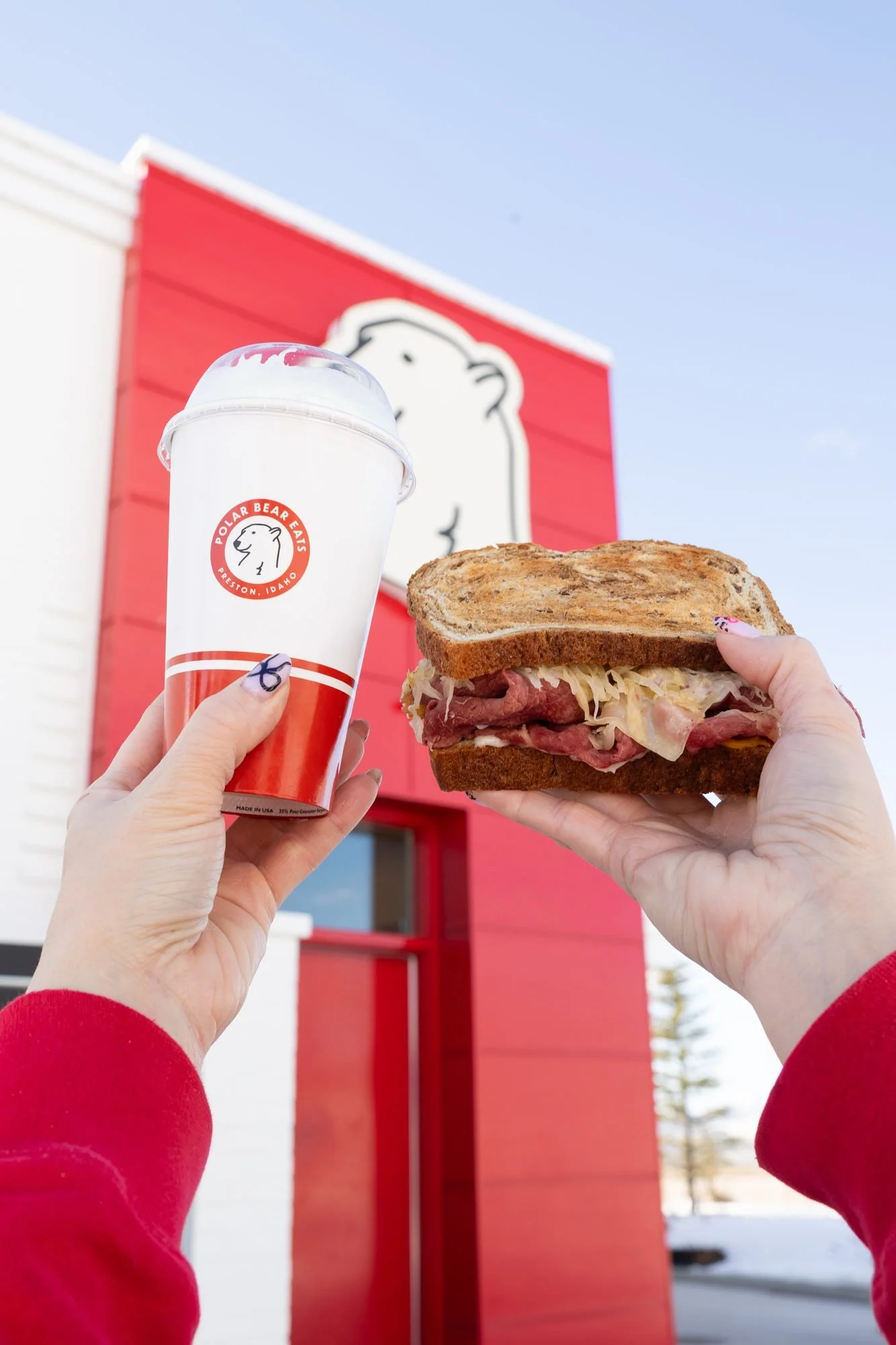 Hands hold up a sandwich and a shake in front of a red and white restaurant building with a polar bear logo