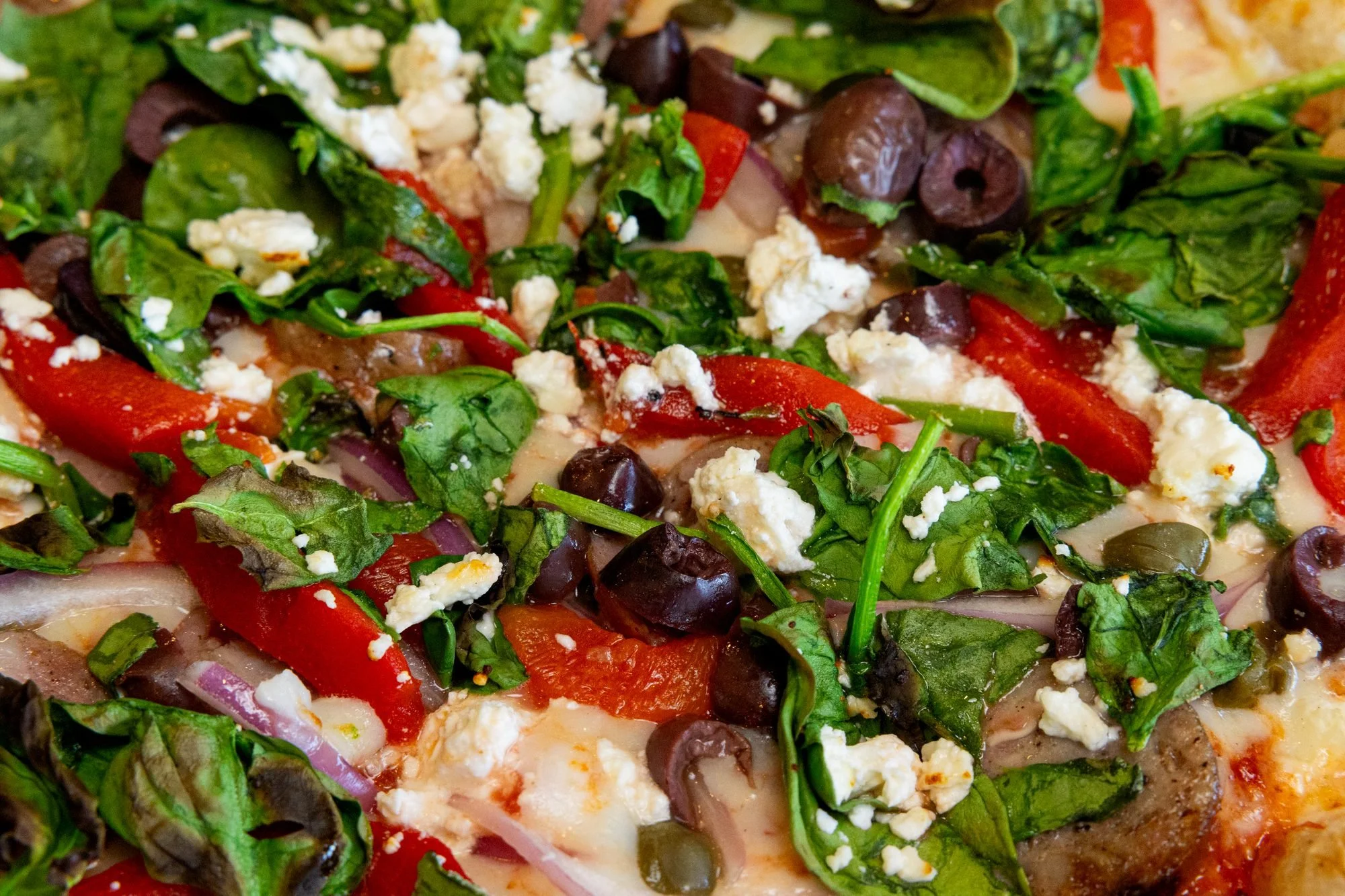 Close view of a wood-fired pizza topped with spinach, roasted red peppers, olives, red onion and feta