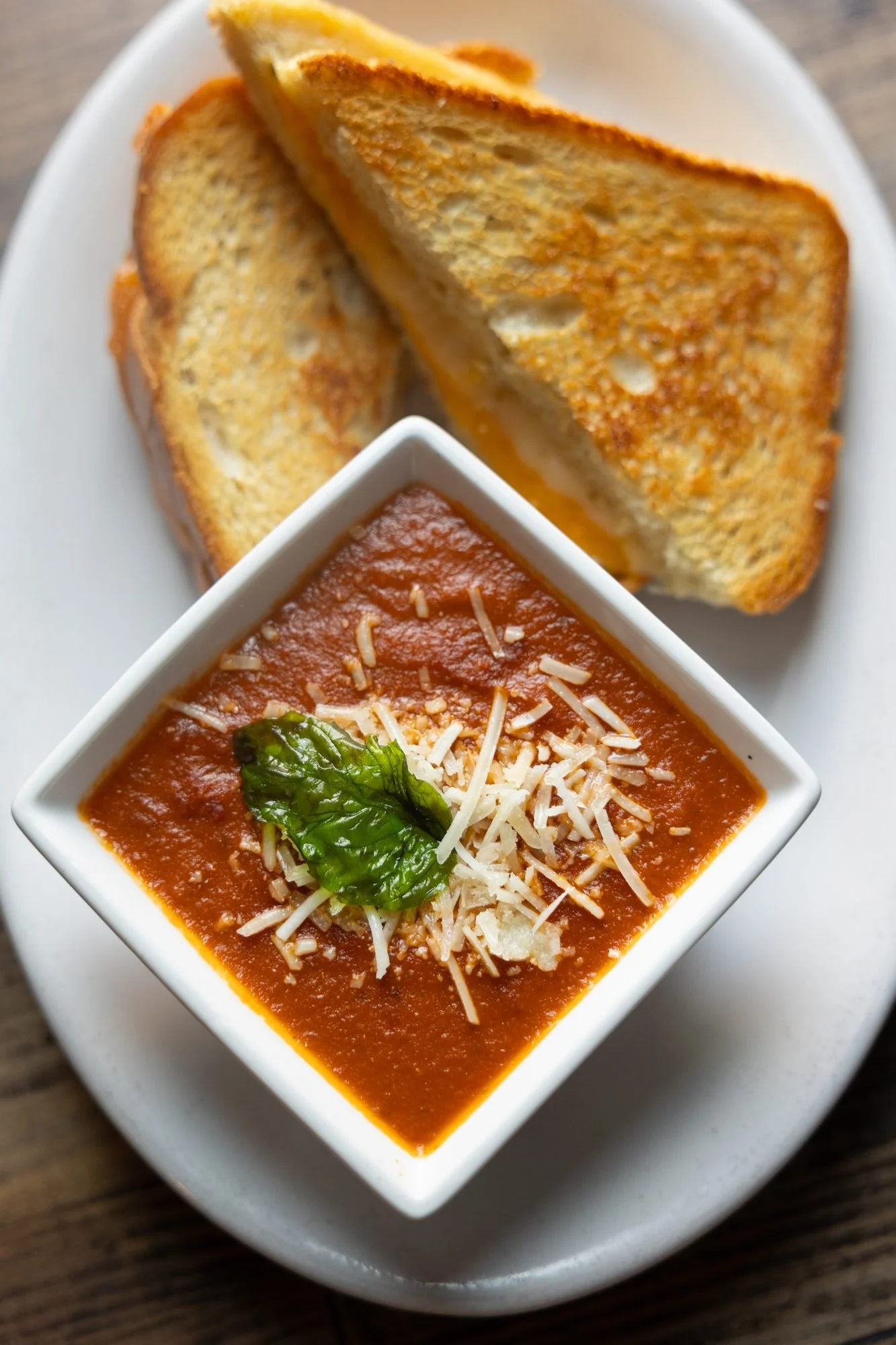 Grilled cheese halves and a square bowl of tomato soup topped with parmesan and basil