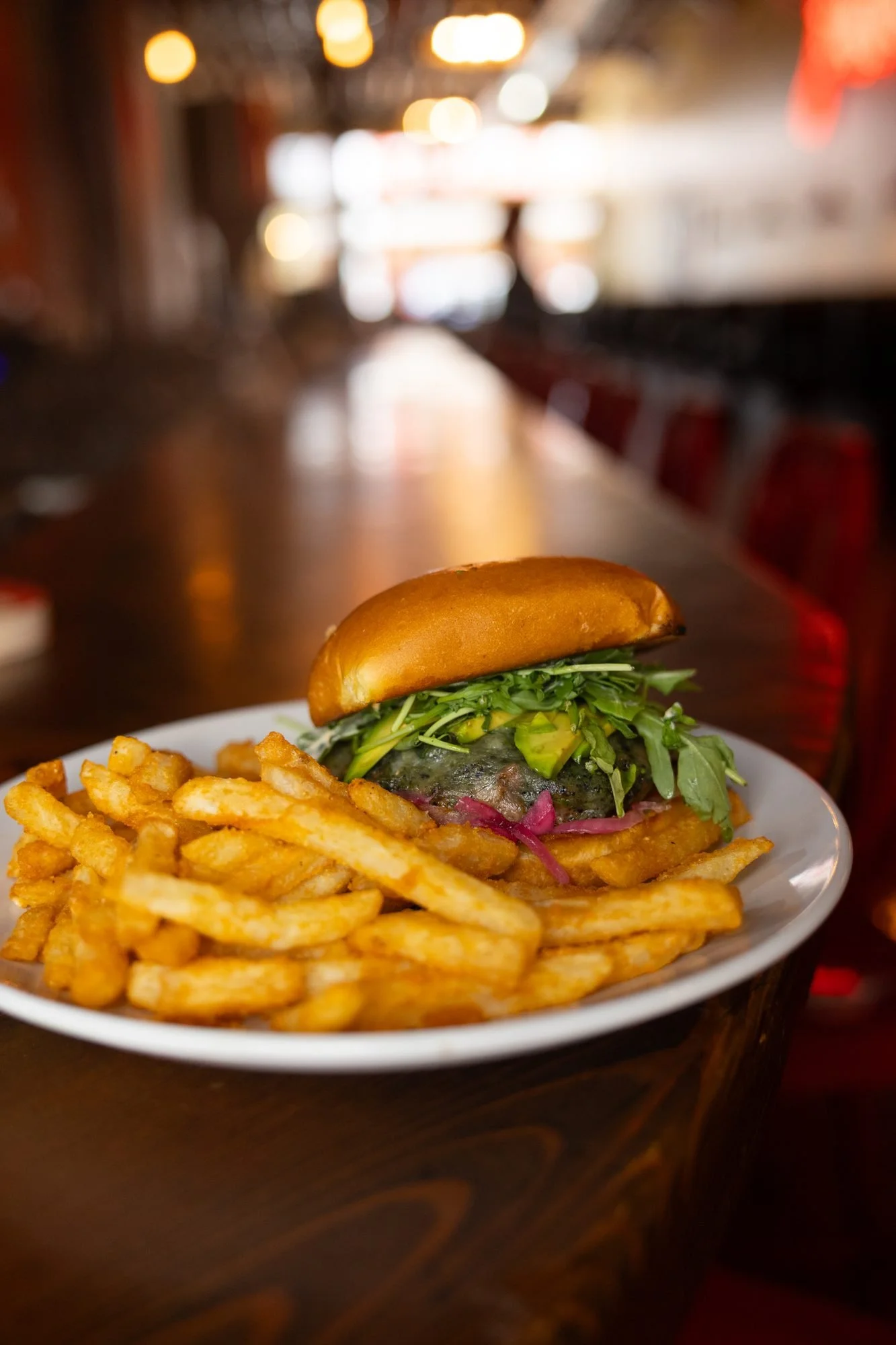 A burger with greens and fries on a white plate on a long wooden bar with warm lights behind