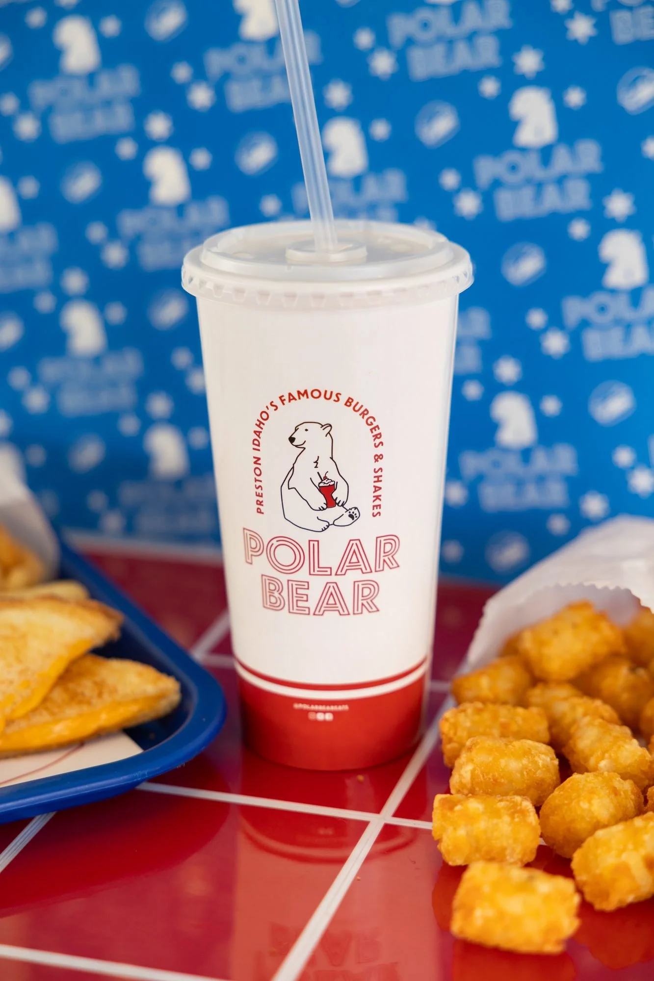 A white Polar Bear cup with a straw on a red tray beside tater tots, against a blue polar bear print backdrop