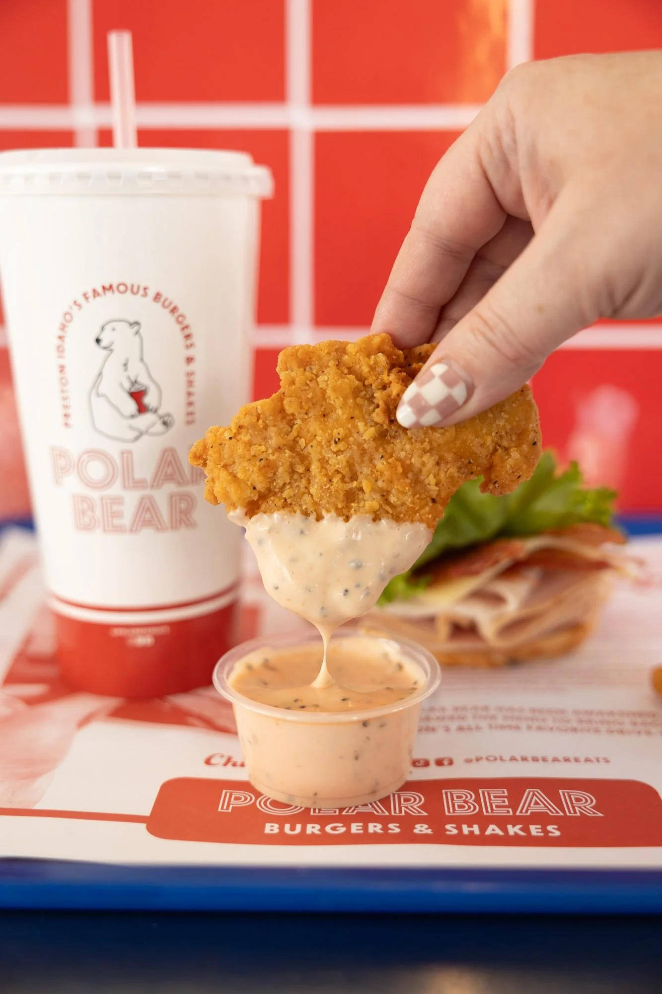 A hand dips a chicken tender into sauce beside a Polar Bear cup and sandwich