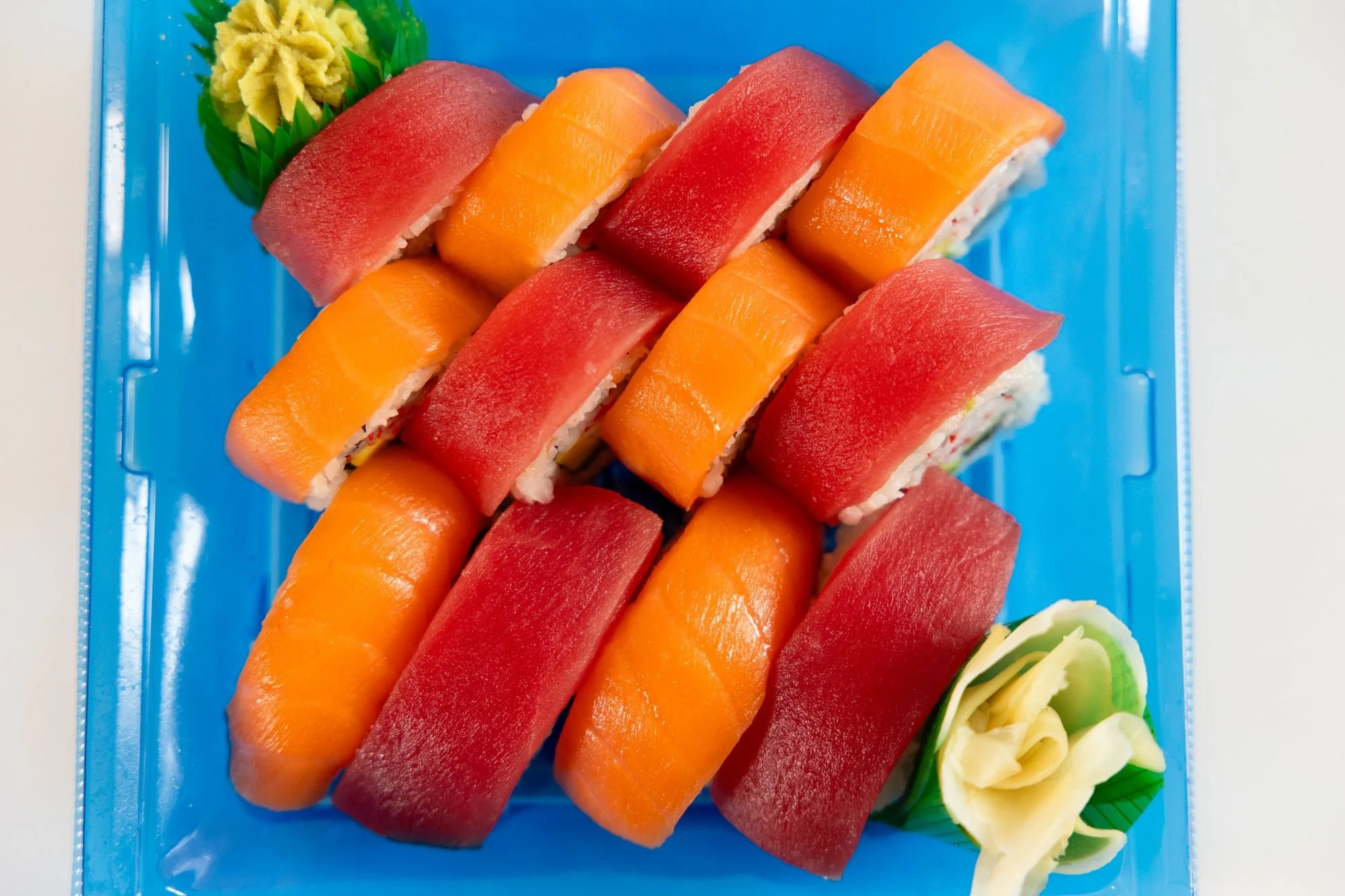 A blue tray of sushi rolls topped with alternating tuna and salmon, with wasabi and pickled ginger