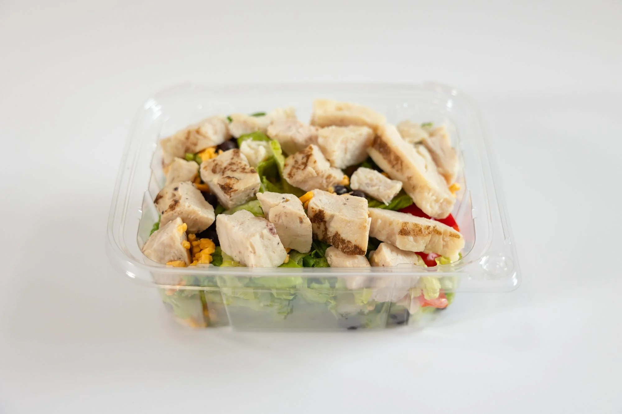 A clear takeout container of chicken salad with corn and greens on a white background