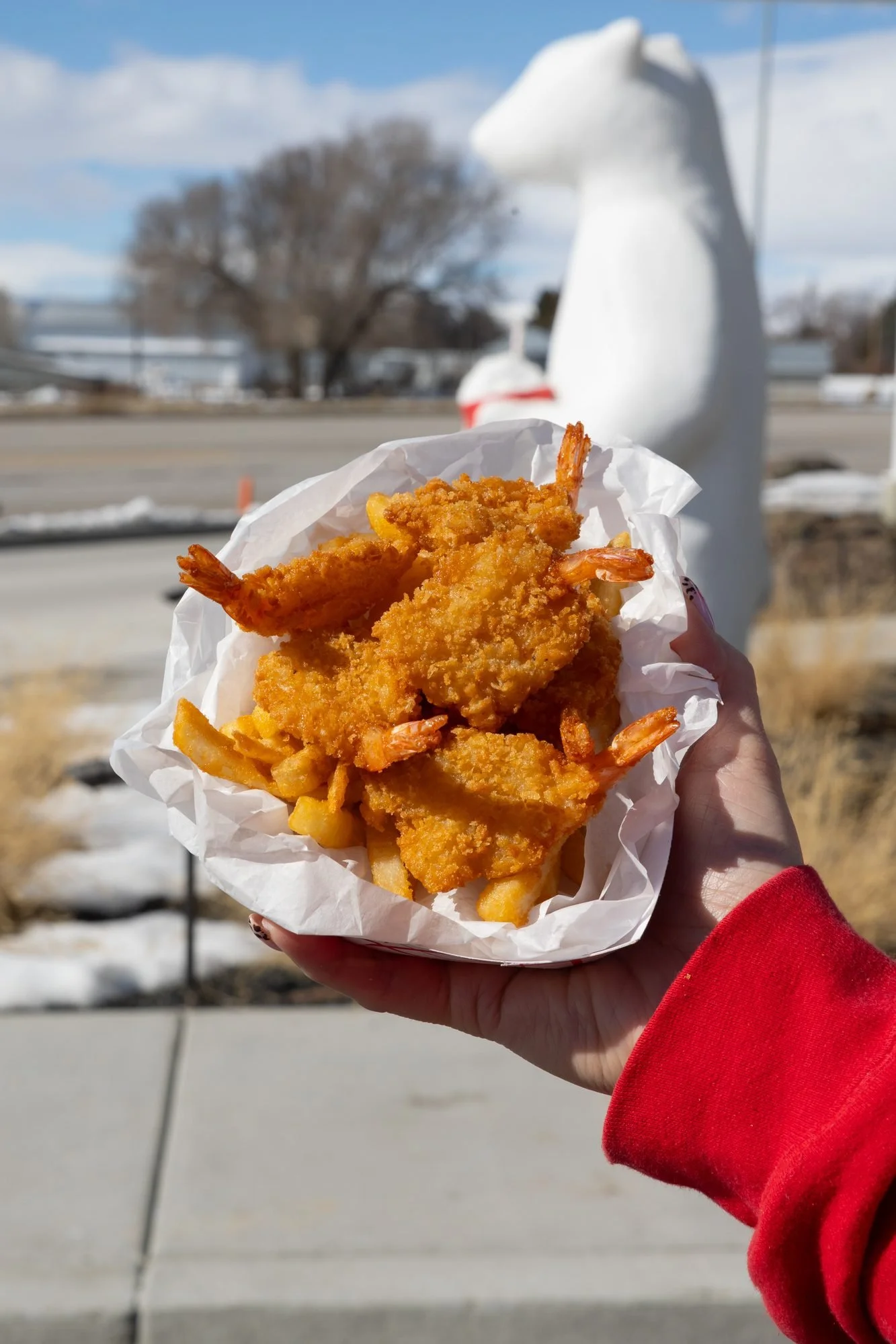 A hand holds a basket of fried shrimp and fries outdoors beside a white polar bear statue with trees behind