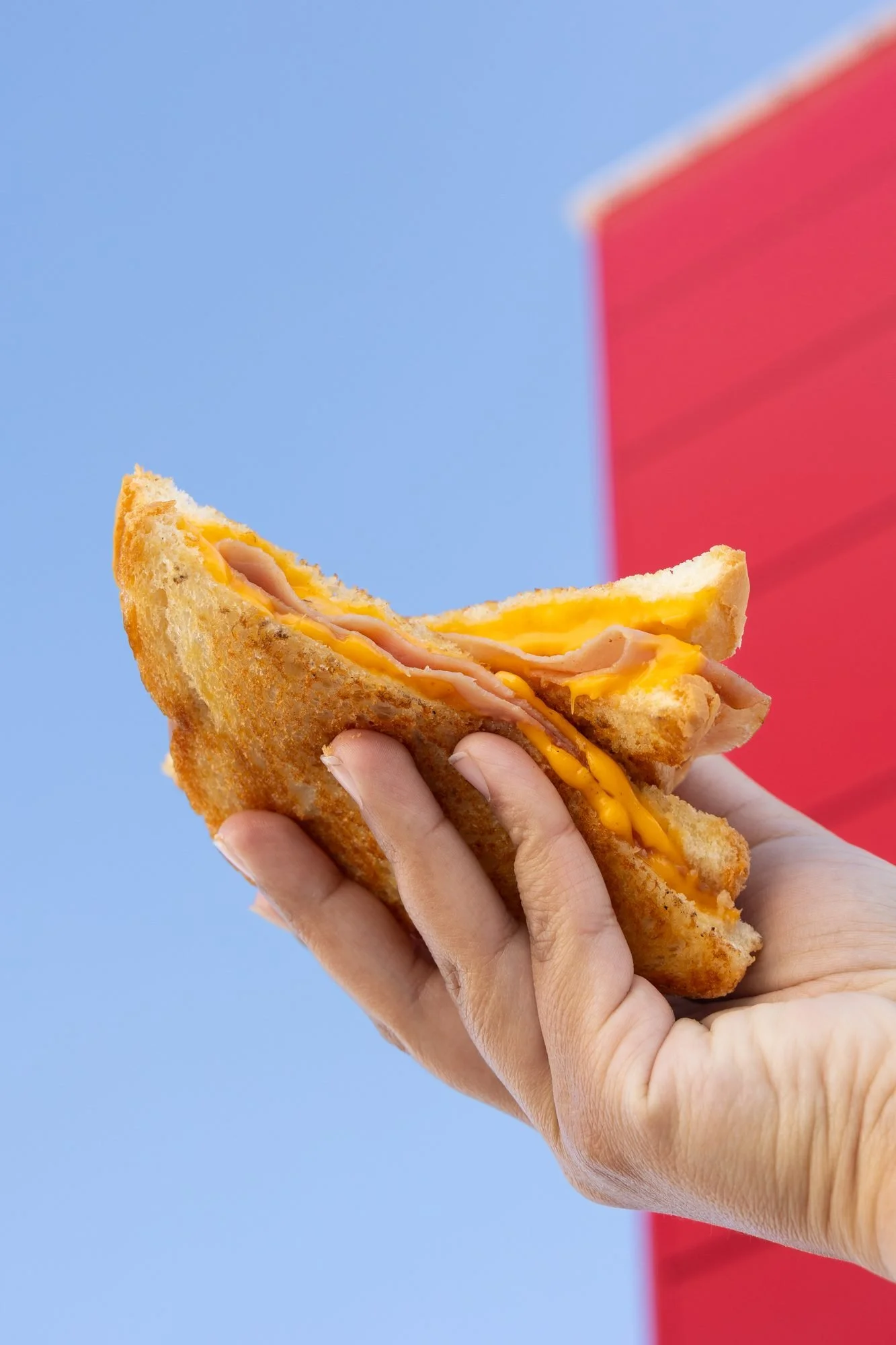 A hand holds a toasted sandwich up against a clear blue sky beside a red building