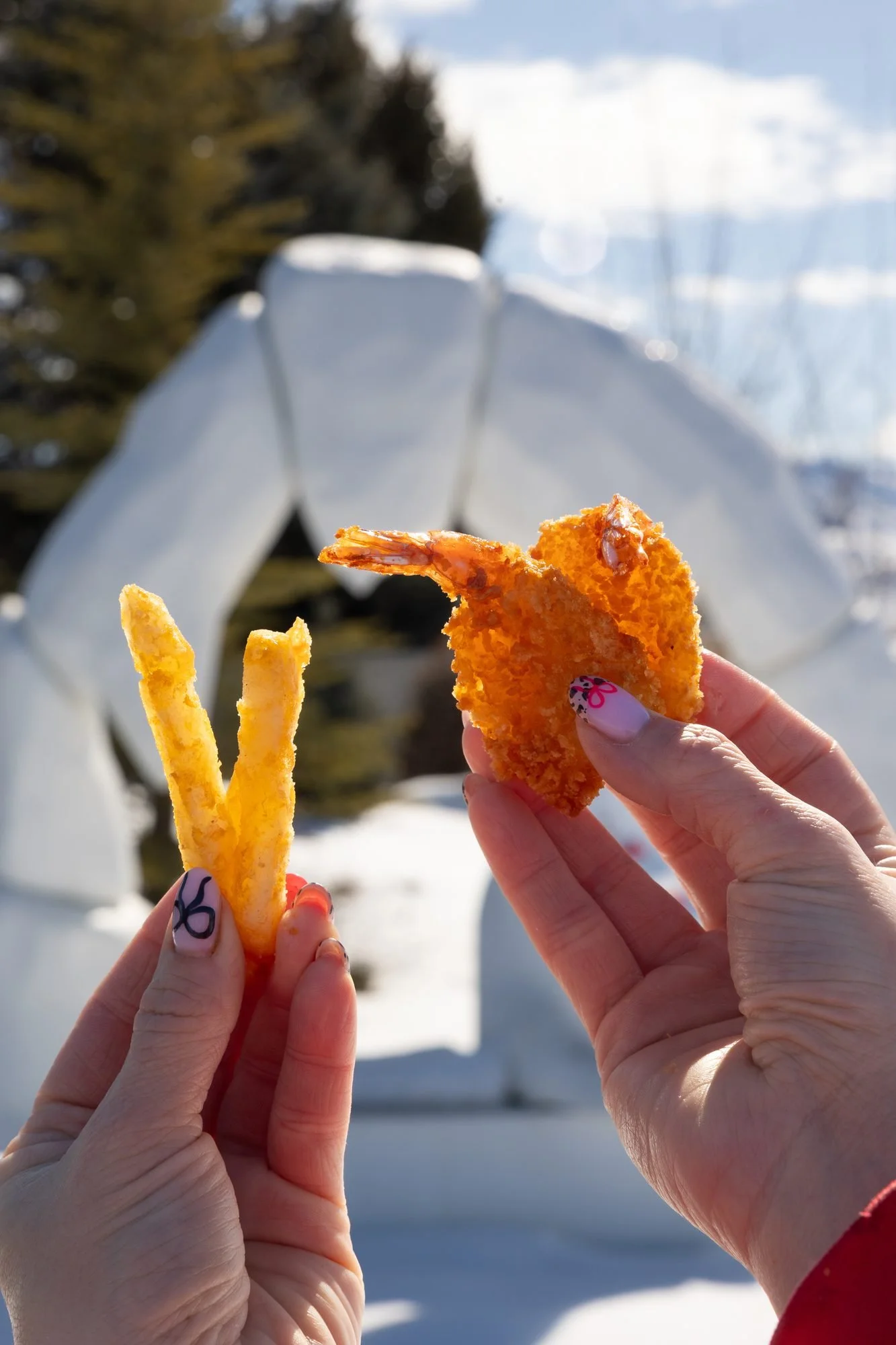 Two hands hold a fry and a fried shrimp in front of a snowy outdoor statue