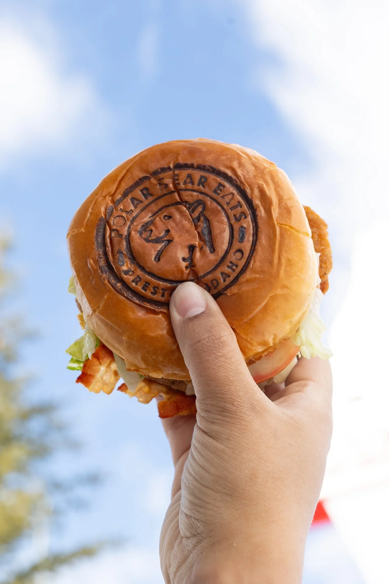 A hand holds a branded burger bun up against a blue sky with white clouds