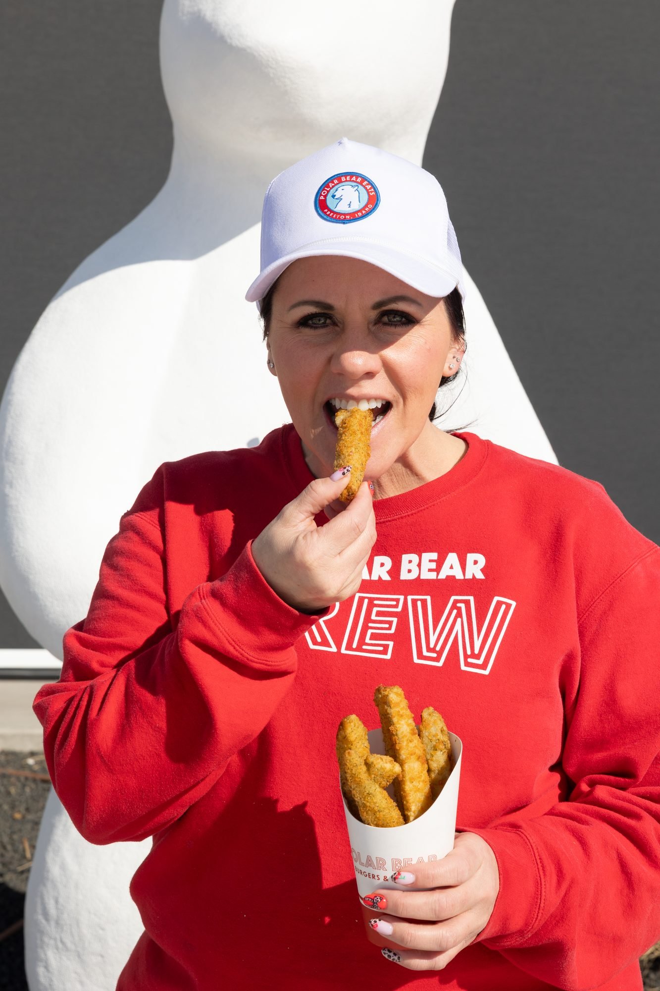 A woman in a red Polar Bear Crew sweatshirt and white cap eats a fry outside beside a polar bear statue