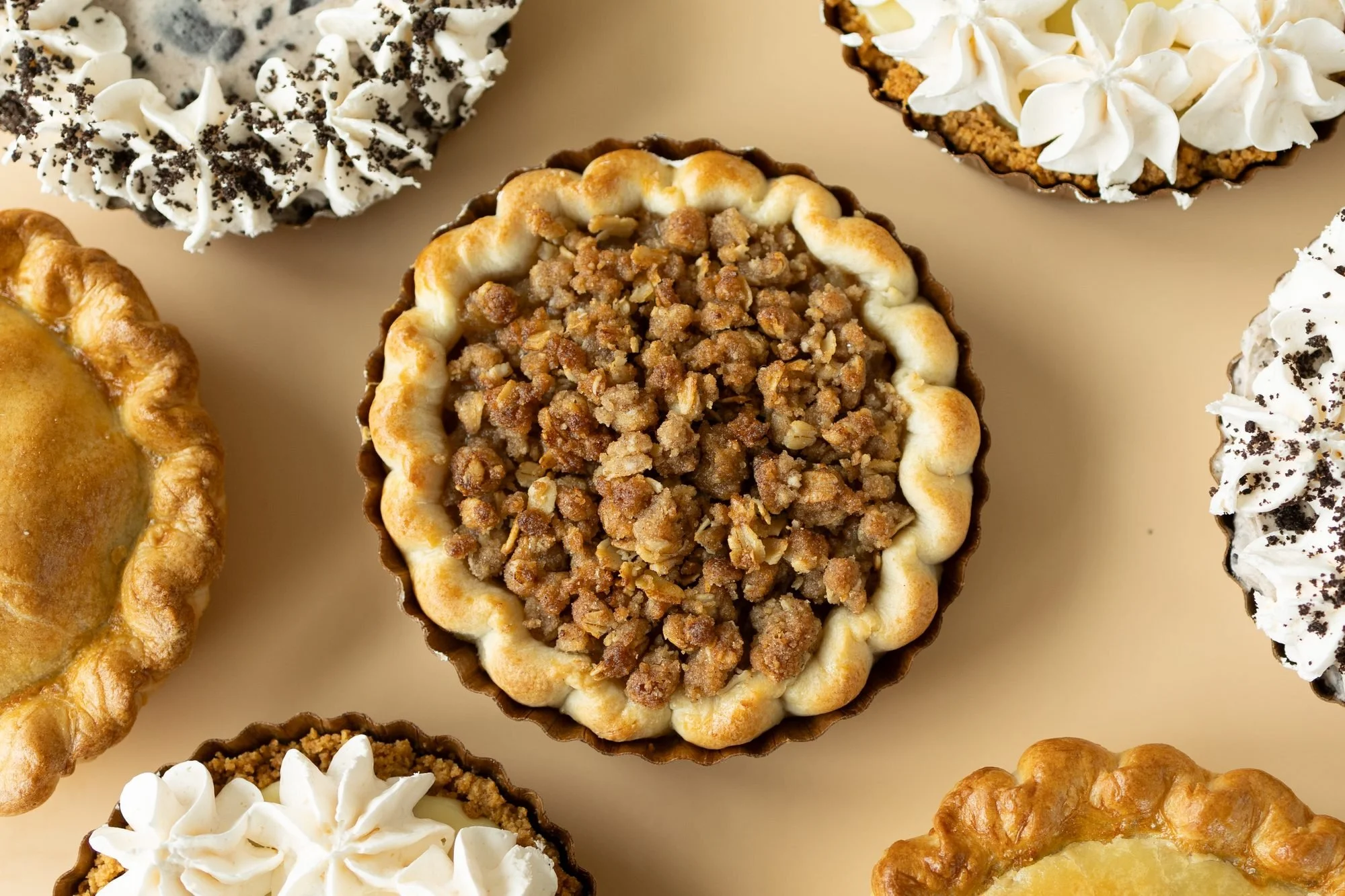 An overhead spread of Crust Club pies with crumble, cookie and whipped cream key lime toppings on a tan background