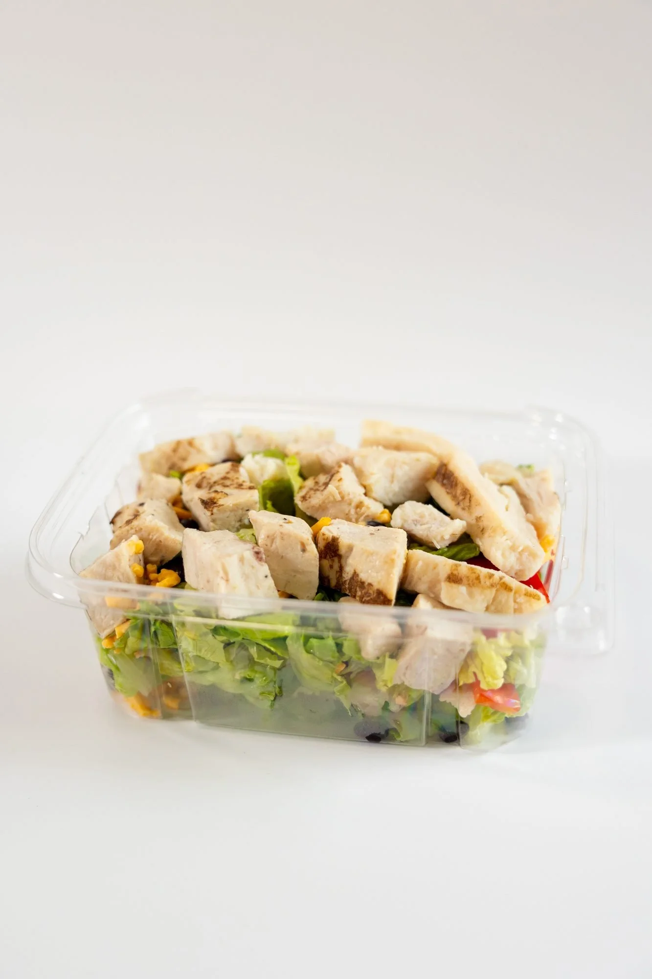 A clear container of grilled chicken salad with corn and greens on a white background