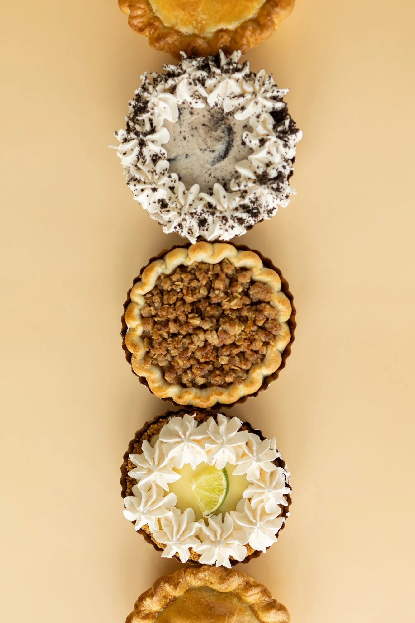 A row of Crust Club pies with cookie, crumble and key lime toppings lined up on a tan background