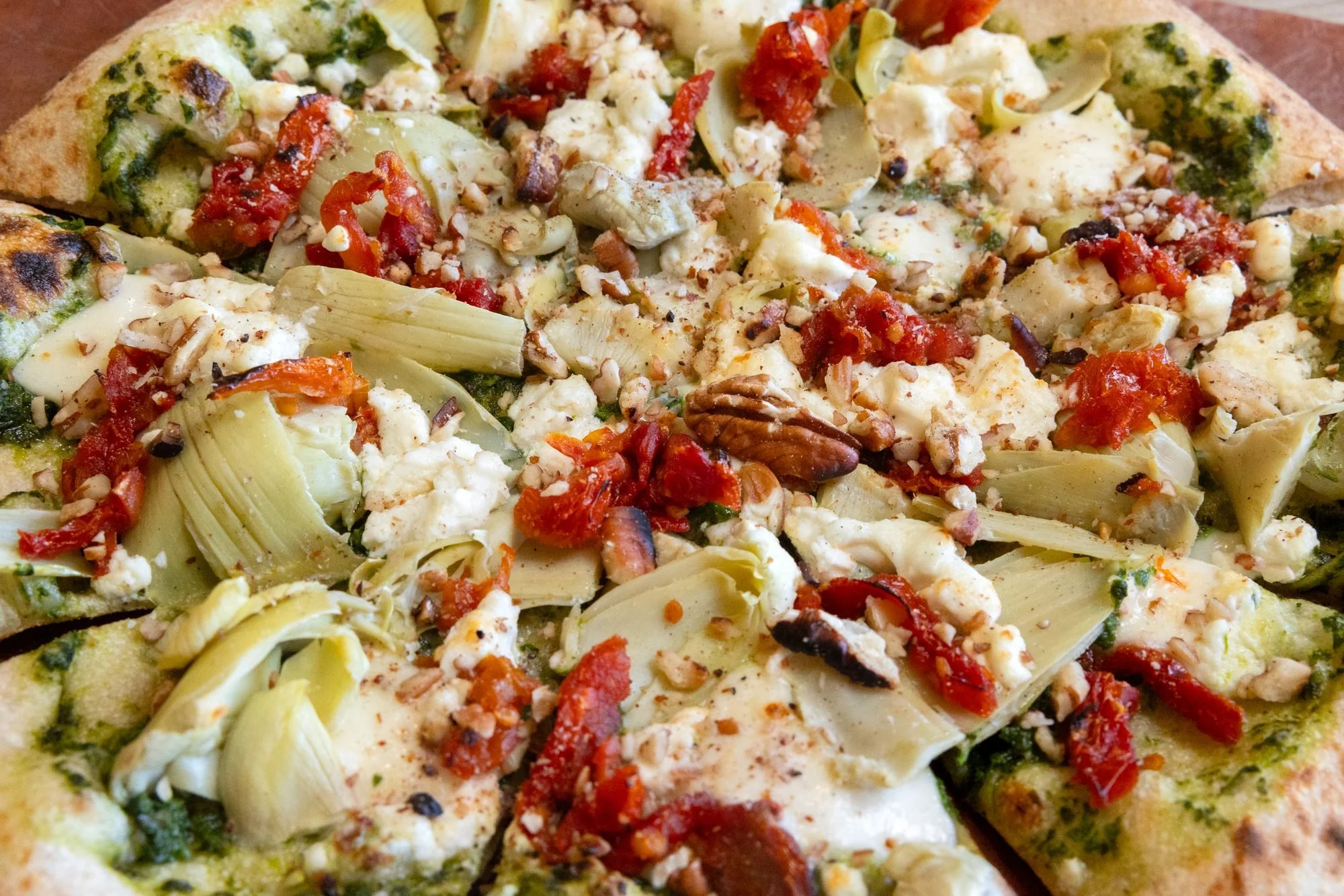 Close view of a wood fired pizza topped with artichoke hearts, sun dried tomatoes, white cheese and pesto