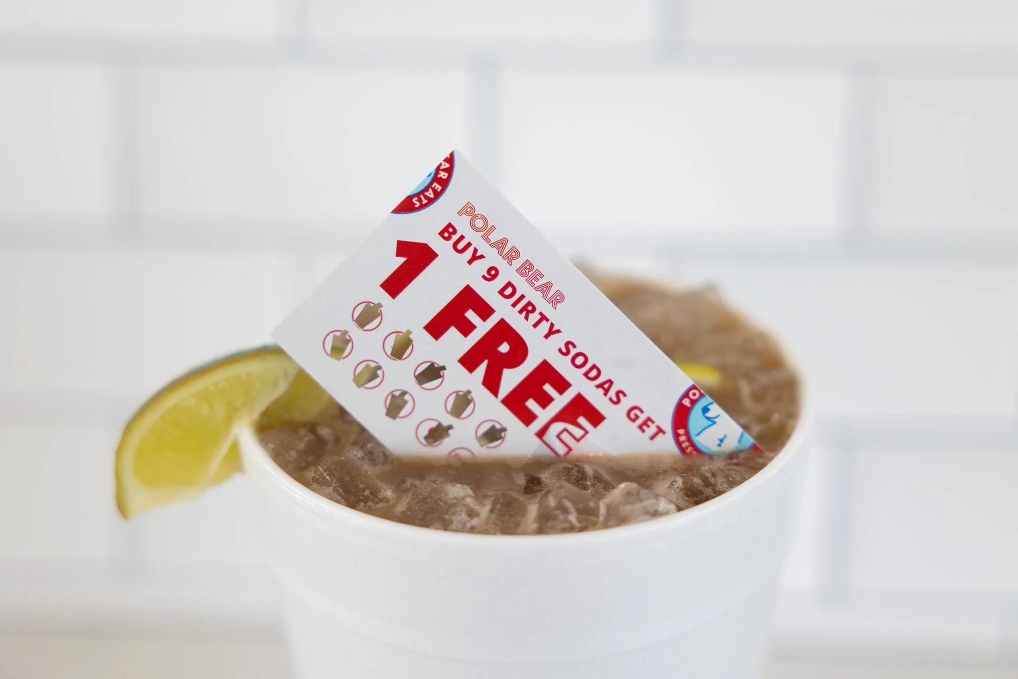 A white foam cup of iced soda with a lime wedge and a Polar Bear punch card reading Buy 9 Dirty Sodas Get 1 Free tucked in the top