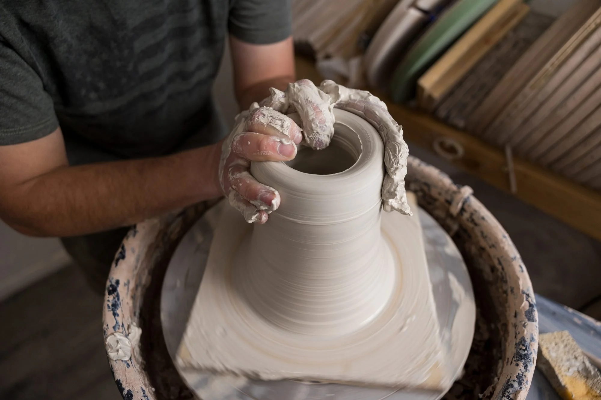 Clay-covered hands pulling up the walls of a clay cylinder on a pottery wheel with shelves behind