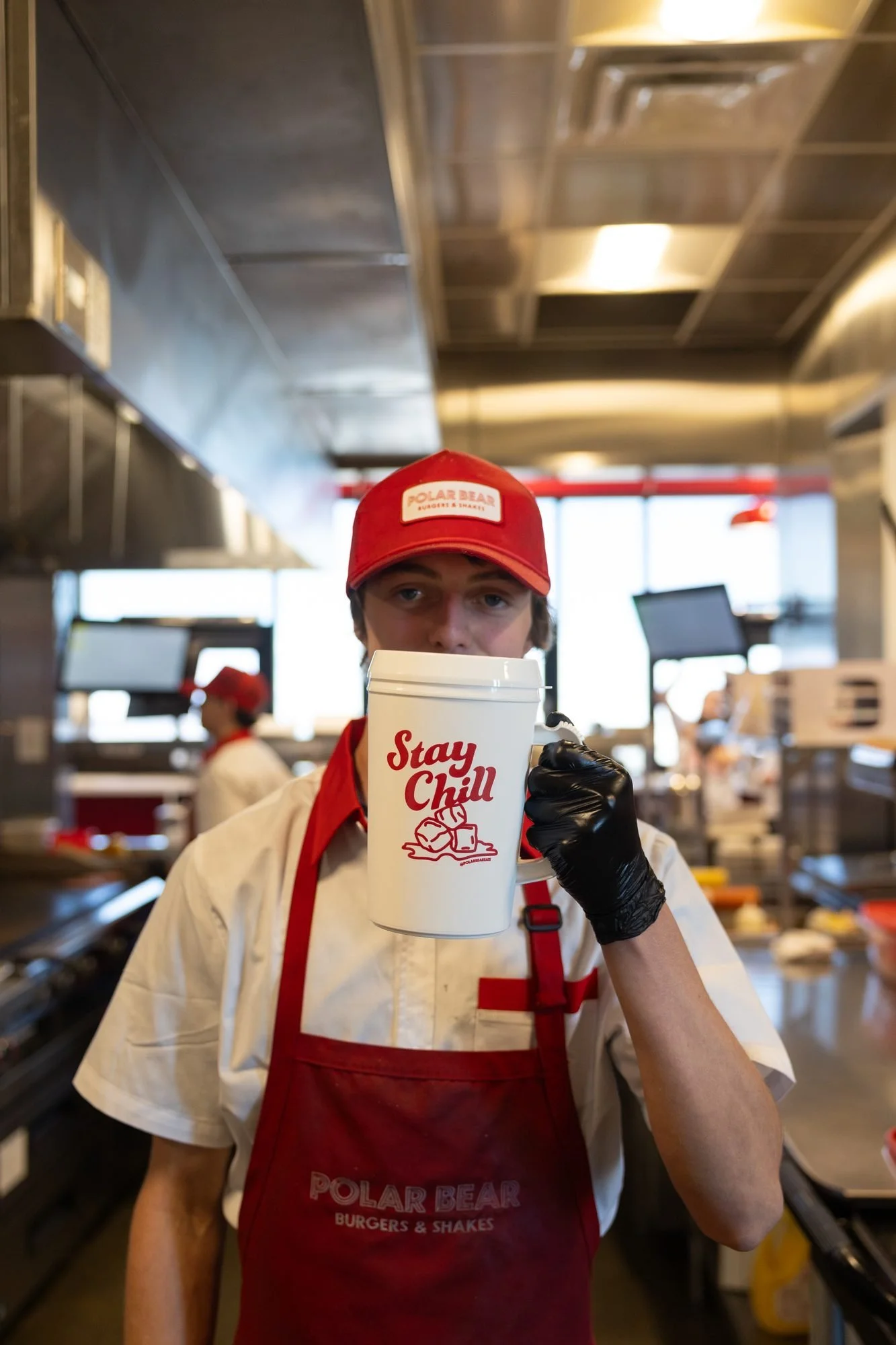 A Polar Bear employee in a red cap and red apron holding a white Stay Chill mug in a restaurant kitchen