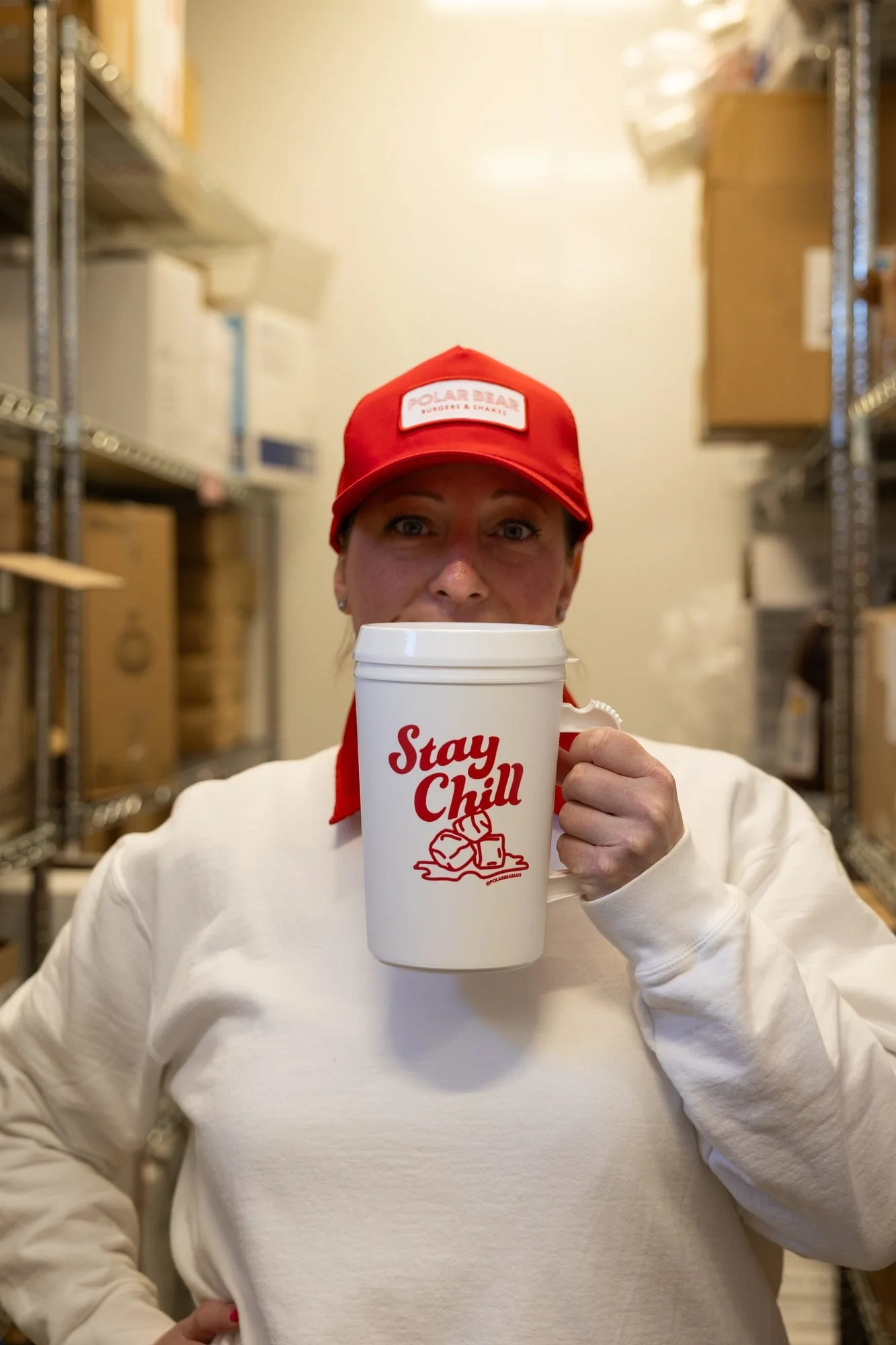 A woman in a red Polar Bear cap holding a white Stay Chill mug in a back room with shelves of supplies